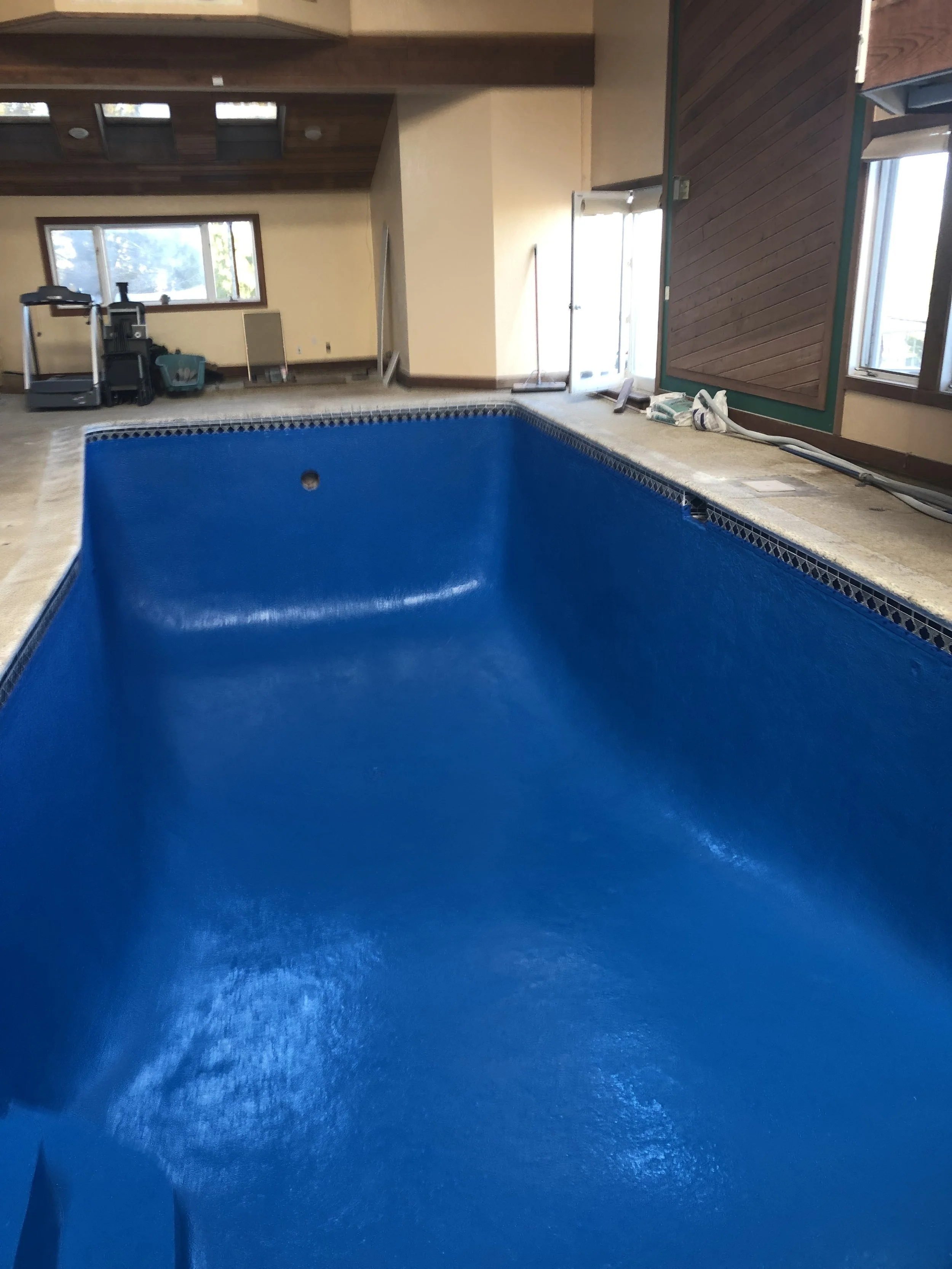Empty indoor pool with a blue finish, under construction or renovation, with tools and construction materials nearby, in a room with windows and wooden ceiling.