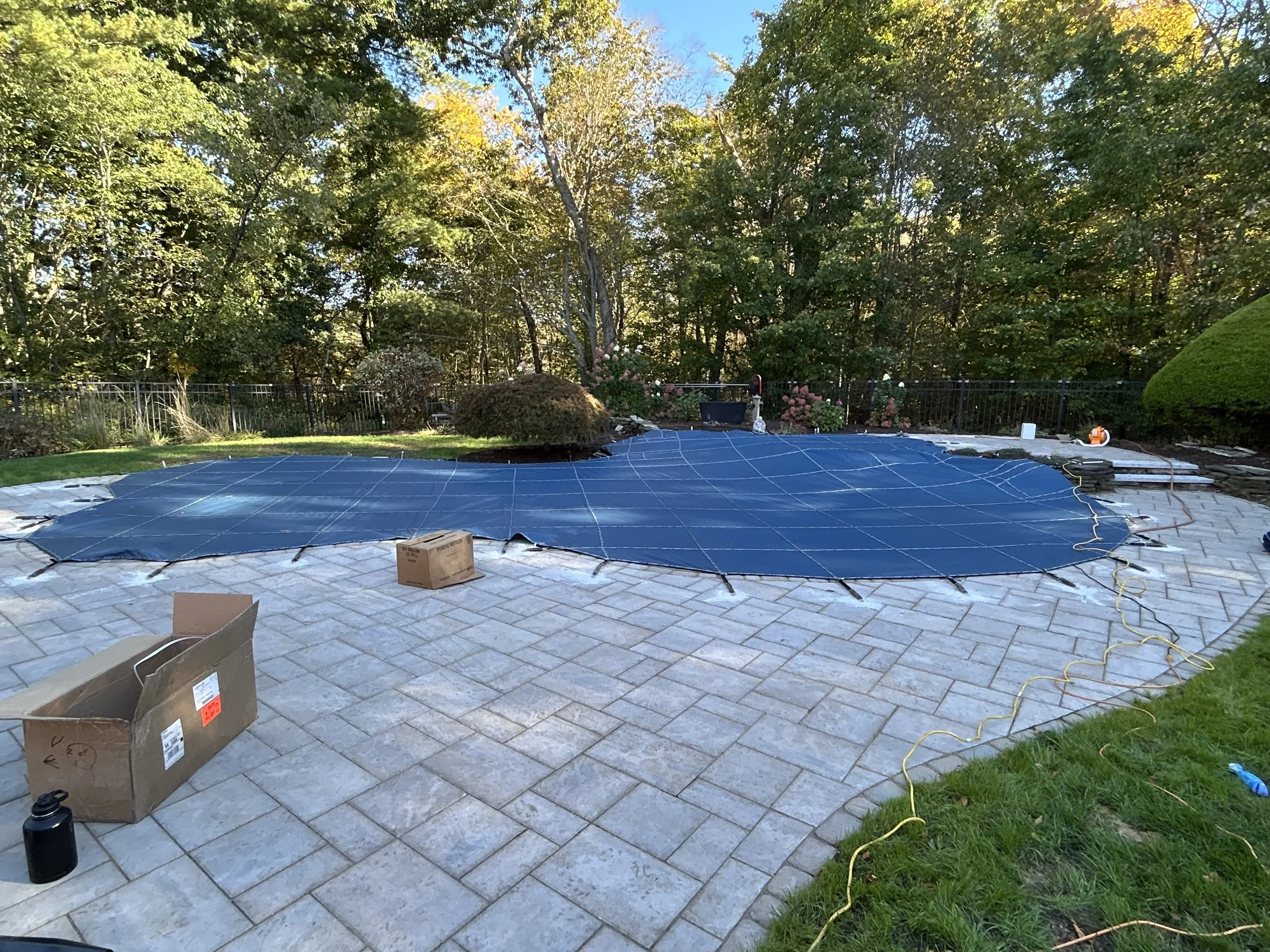 In-ground swimming pool surrounded by a paved patio, with a black safety cover partially rolled out over the pool. There are boxes and tools nearby, and the backyard is enclosed with a black fence, with trees and shrubbery in the background.