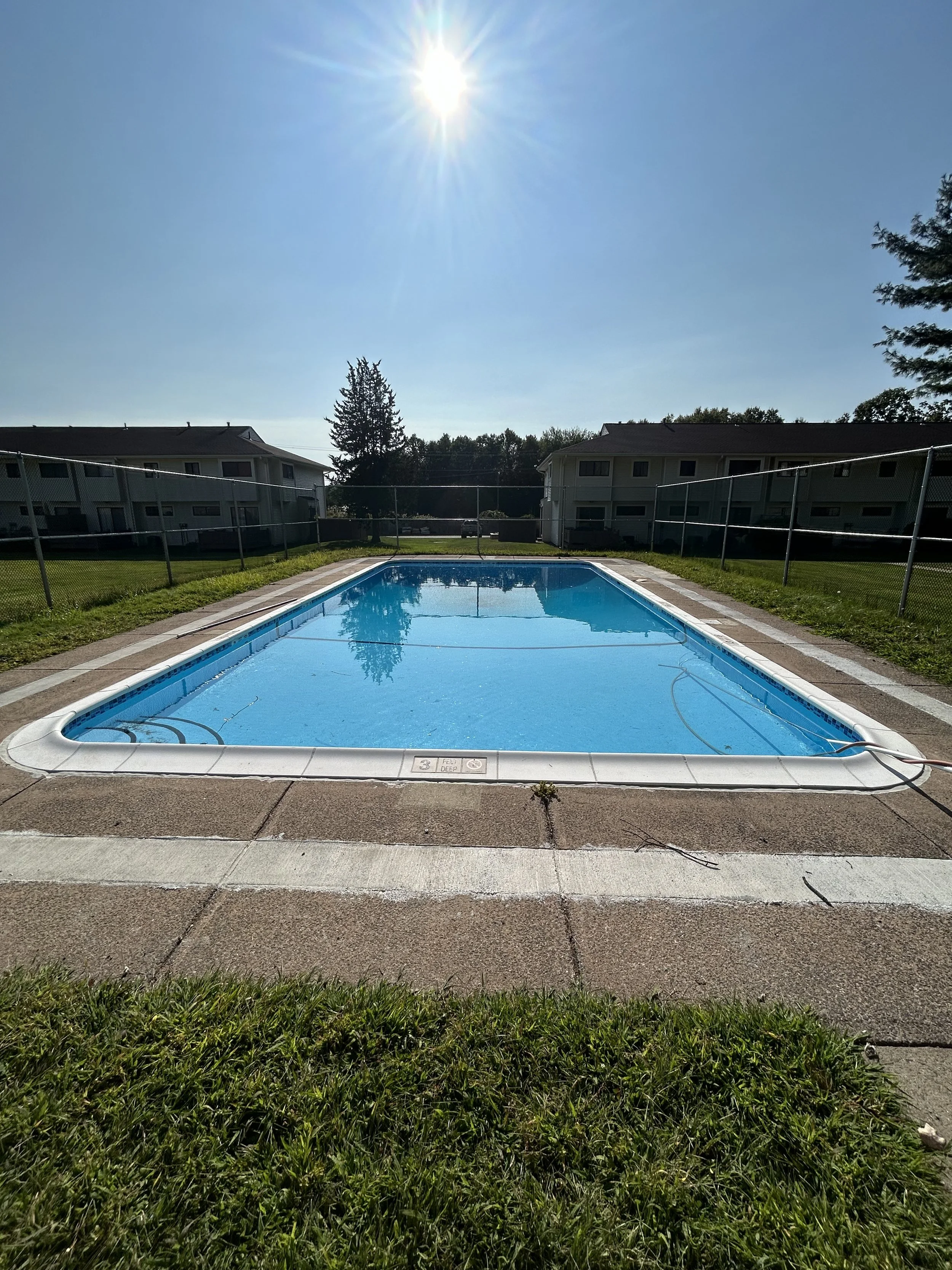Empty swimming pool on a sunny day with a high fence, green grass, and residential buildings in the background.