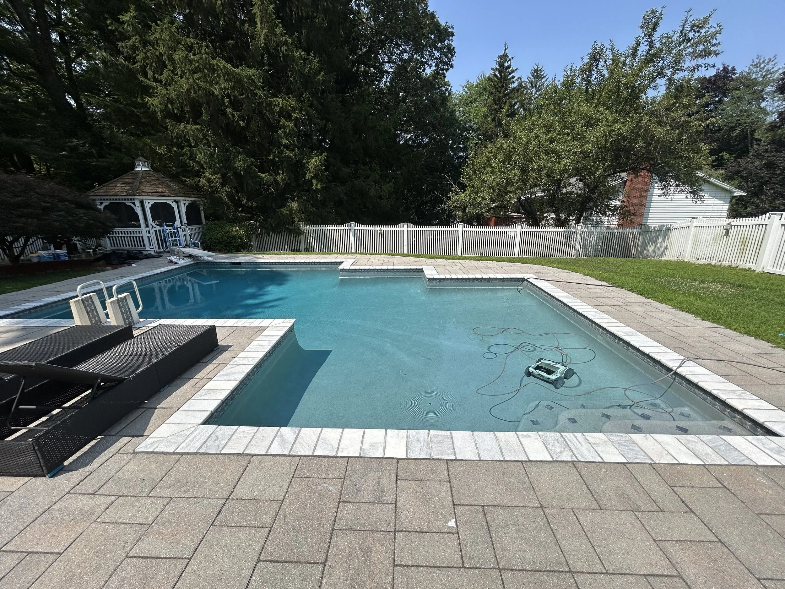 A backyard swimming pool with a white fence, lounge chairs, and a gazebo in the background, surrounded by trees and green grass.