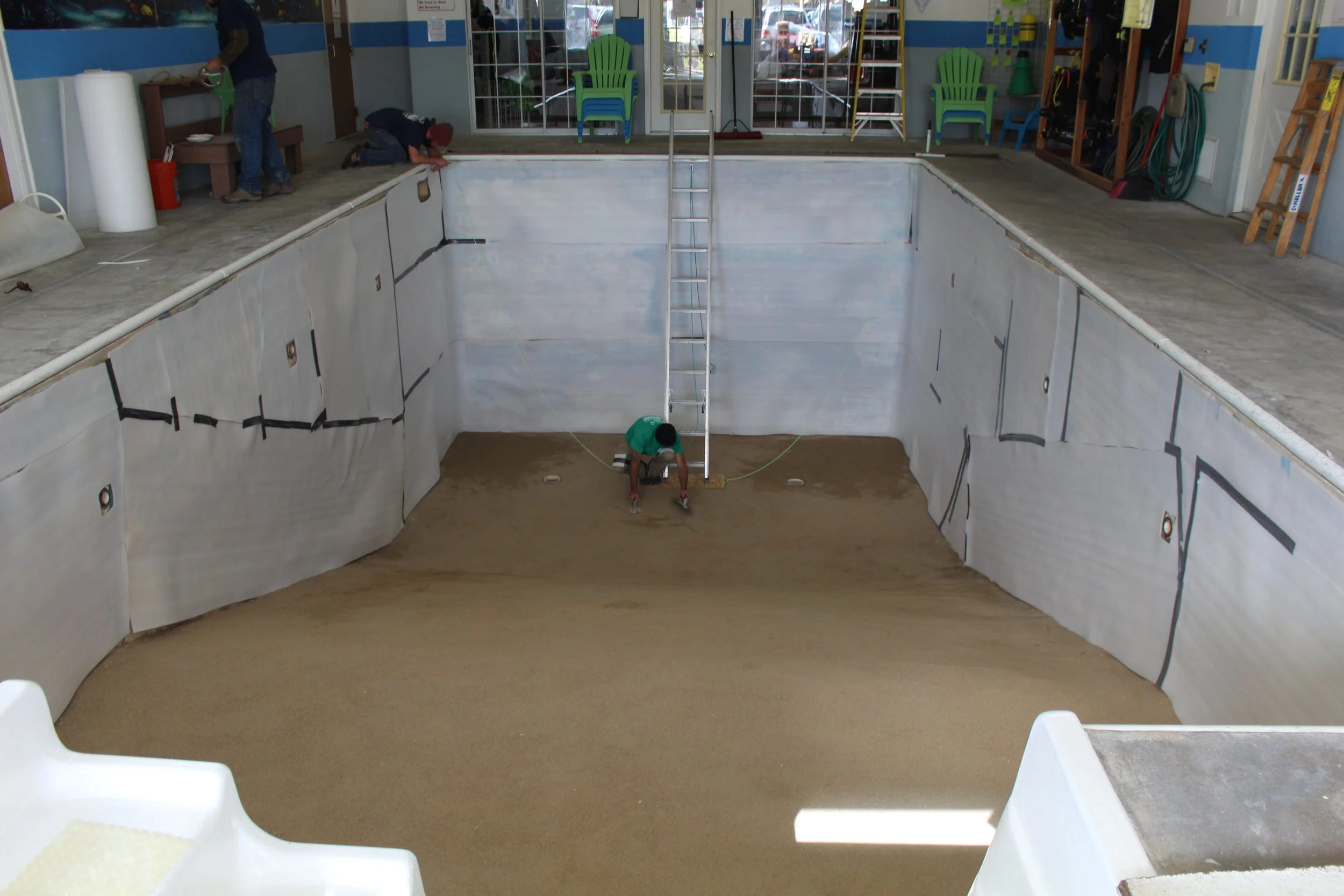 An indoor swimming pool under construction, with a worker preparing the bottom surface and another worker on the side. The pool is empty with a ladder in the center, and walls lined with insulation material. There are chairs and tools stored along th