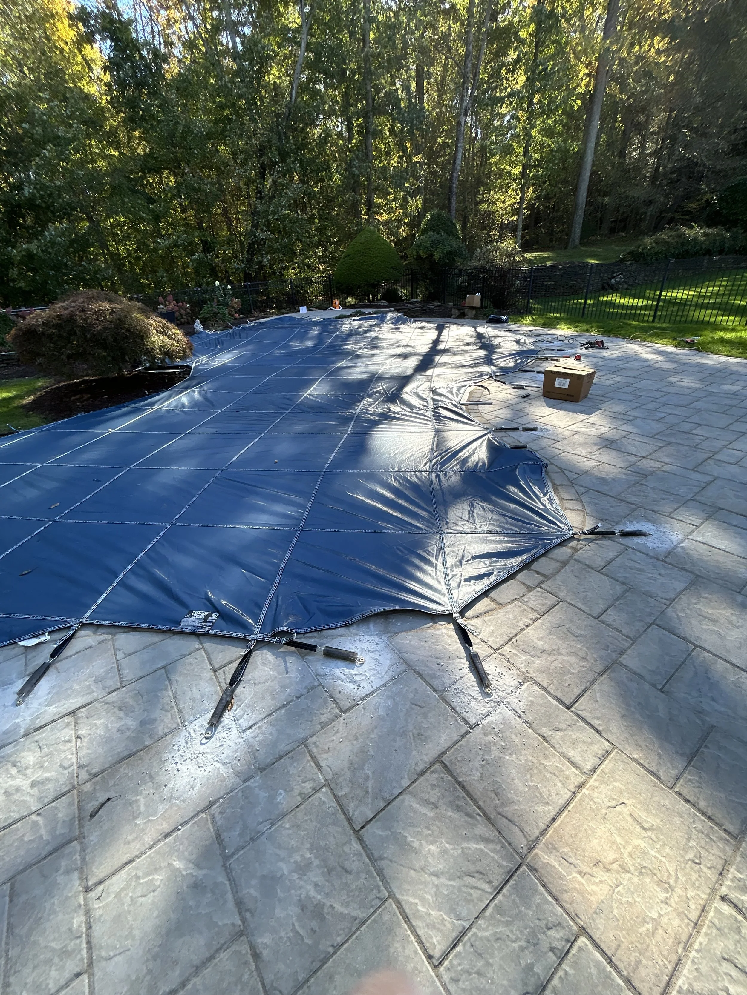 A backyard with a rectangular swimming pool covered with a black winter tarp, secured with straps and stakes. Surrounding the pool are stone tiles, with trees and bushes in the background, and shadows cast by the trees on the tarp.