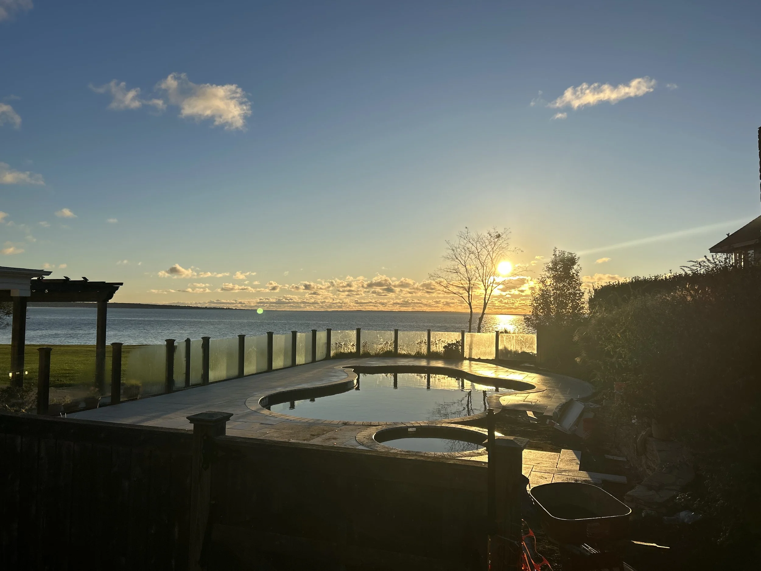 Sunset view over a body of water with a backyard pool in the foreground, surrounded by a wooden fence and trees.