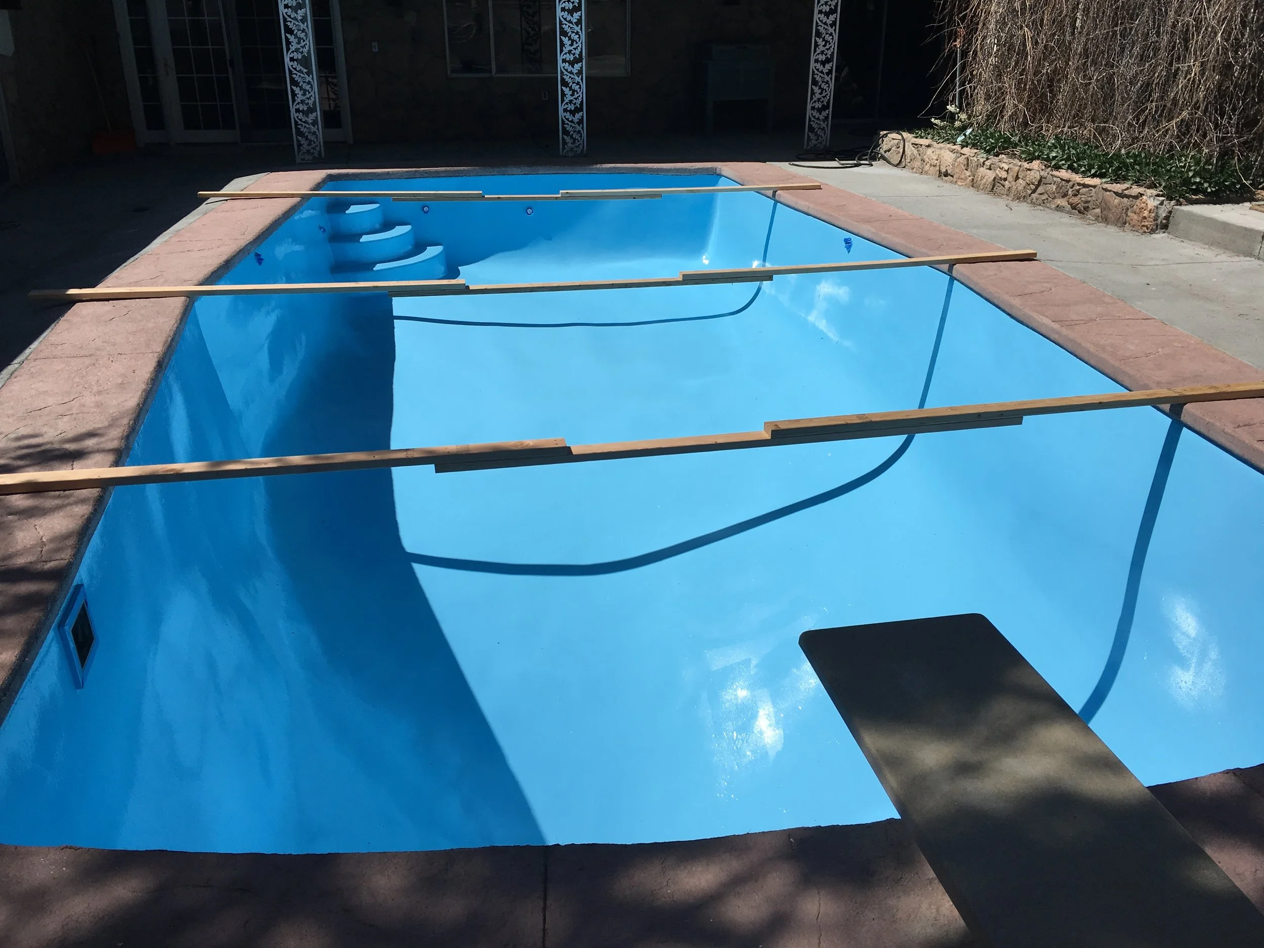 Empty backyard swimming pool with a ladder, surrounded by a concrete patio and construction wooden beams across it, with some shadows of trees on the ground.