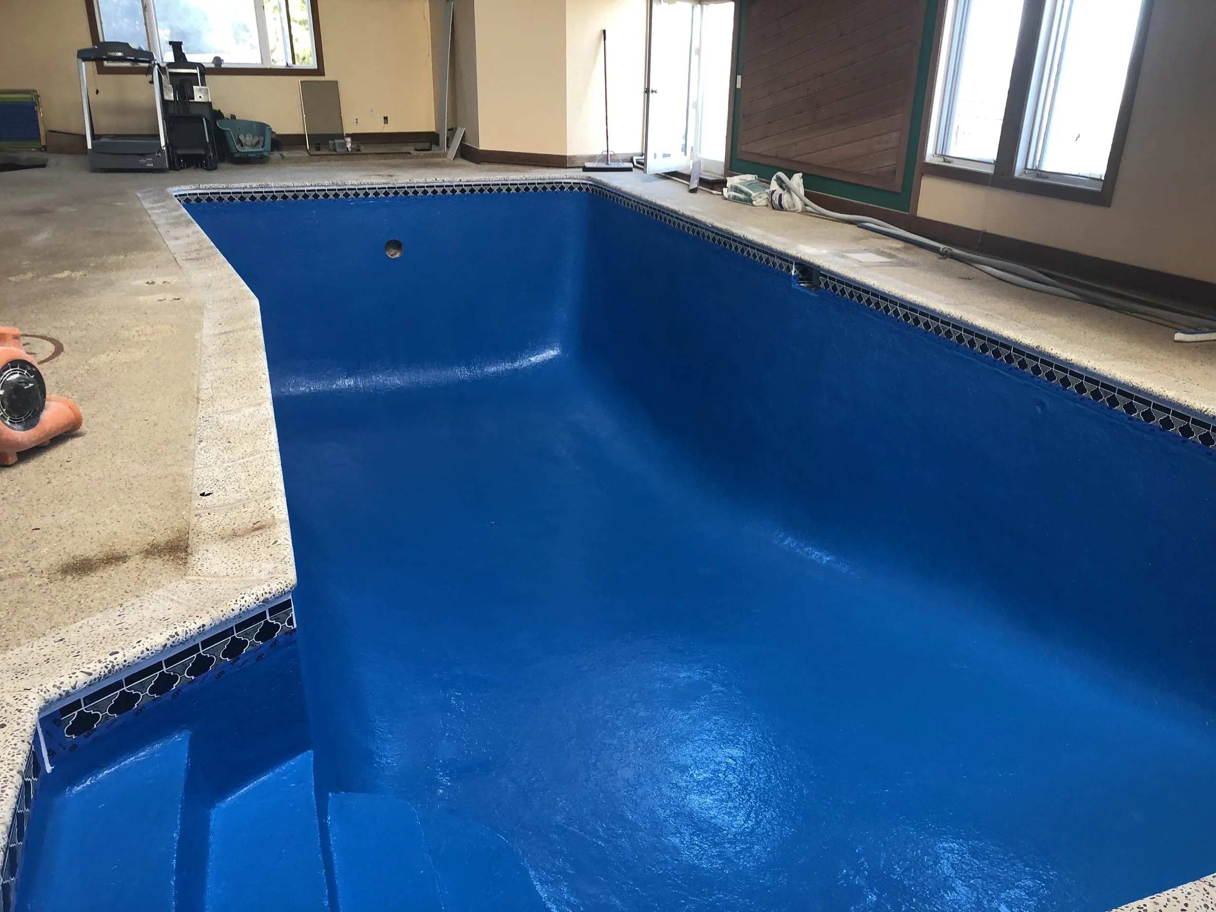 Indoor swimming pool under construction with blue waterproof coating, surrounded by a beige concrete deck, construction tools and supplies on the deck, windows and doors in the background.
