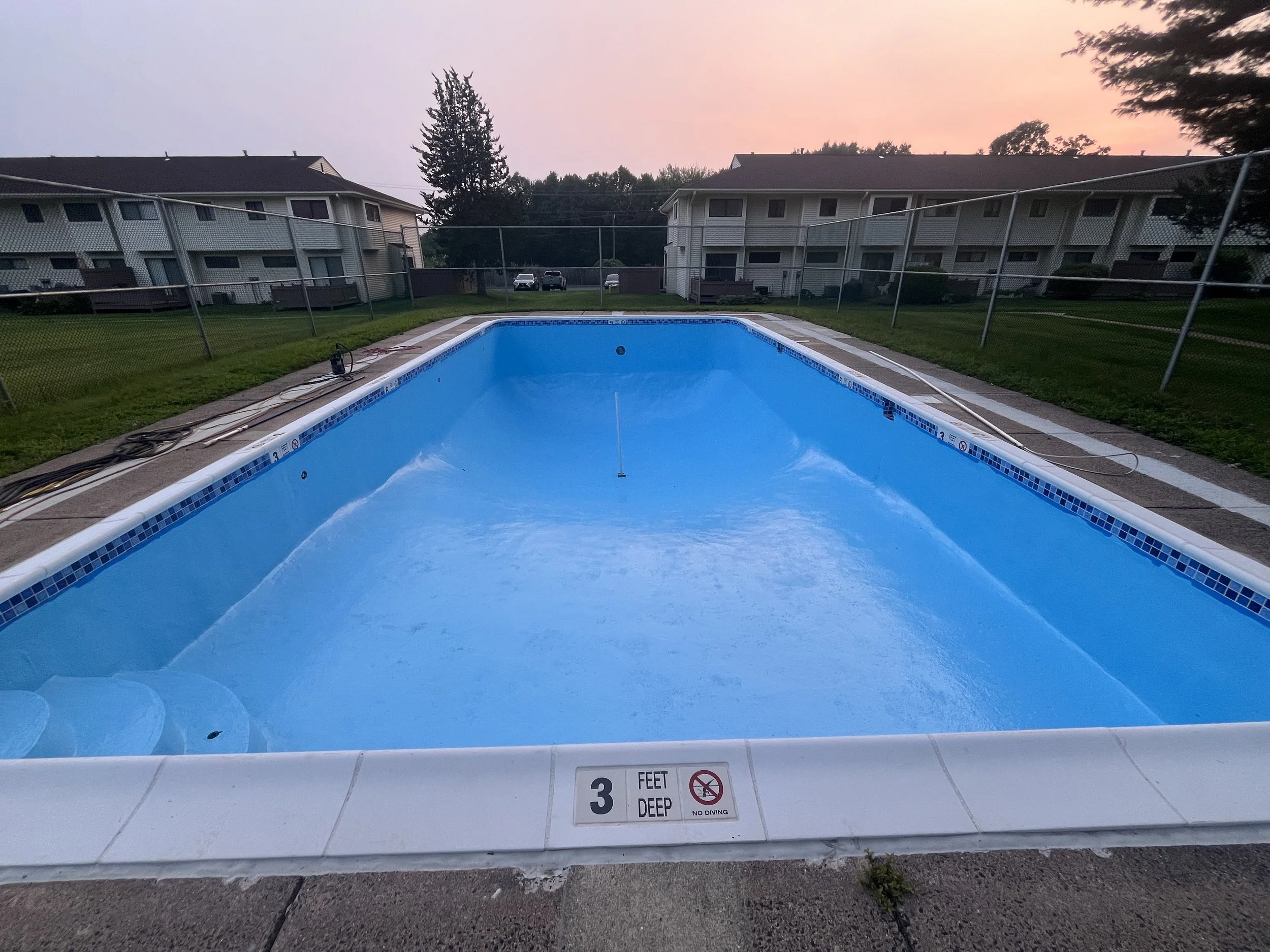 Empty swimming pool with a depth of 3 feet, surrounded by a concrete deck, fences, and residential apartments in the background during sunset.