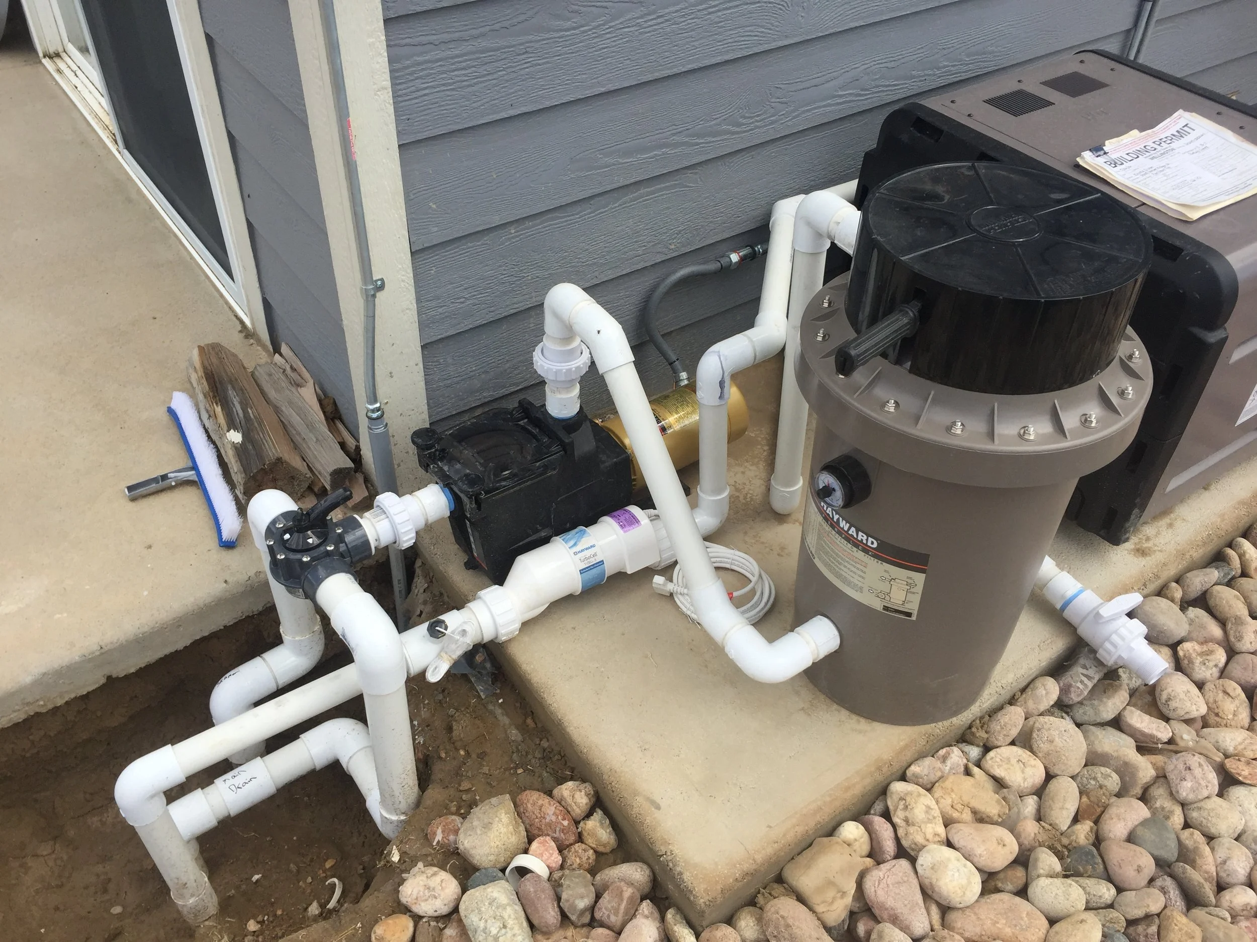 Pool filter system with pipes connected outside a house, next to a concrete slab and rocks.