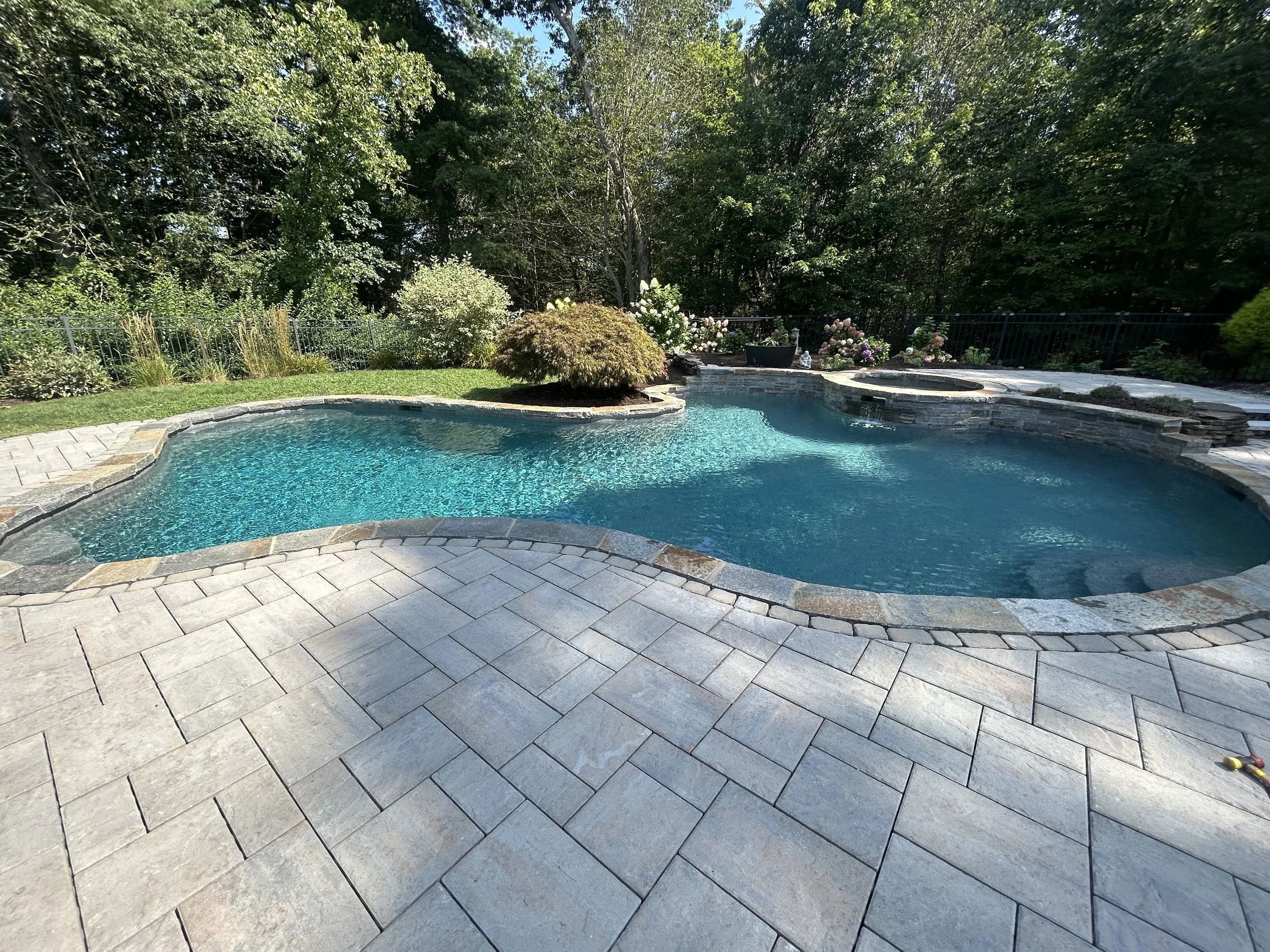 A backyard with a kidney-shaped swimming pool surrounded by a stone patio, lush trees, and flower bushes.