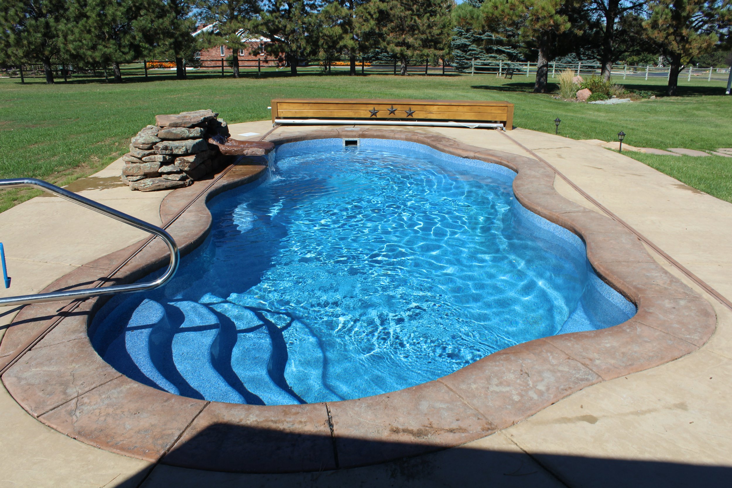 An outdoor swimming pool with a kidney shape, surrounded by a concrete deck and green lawn, with trees and a fence in the background.