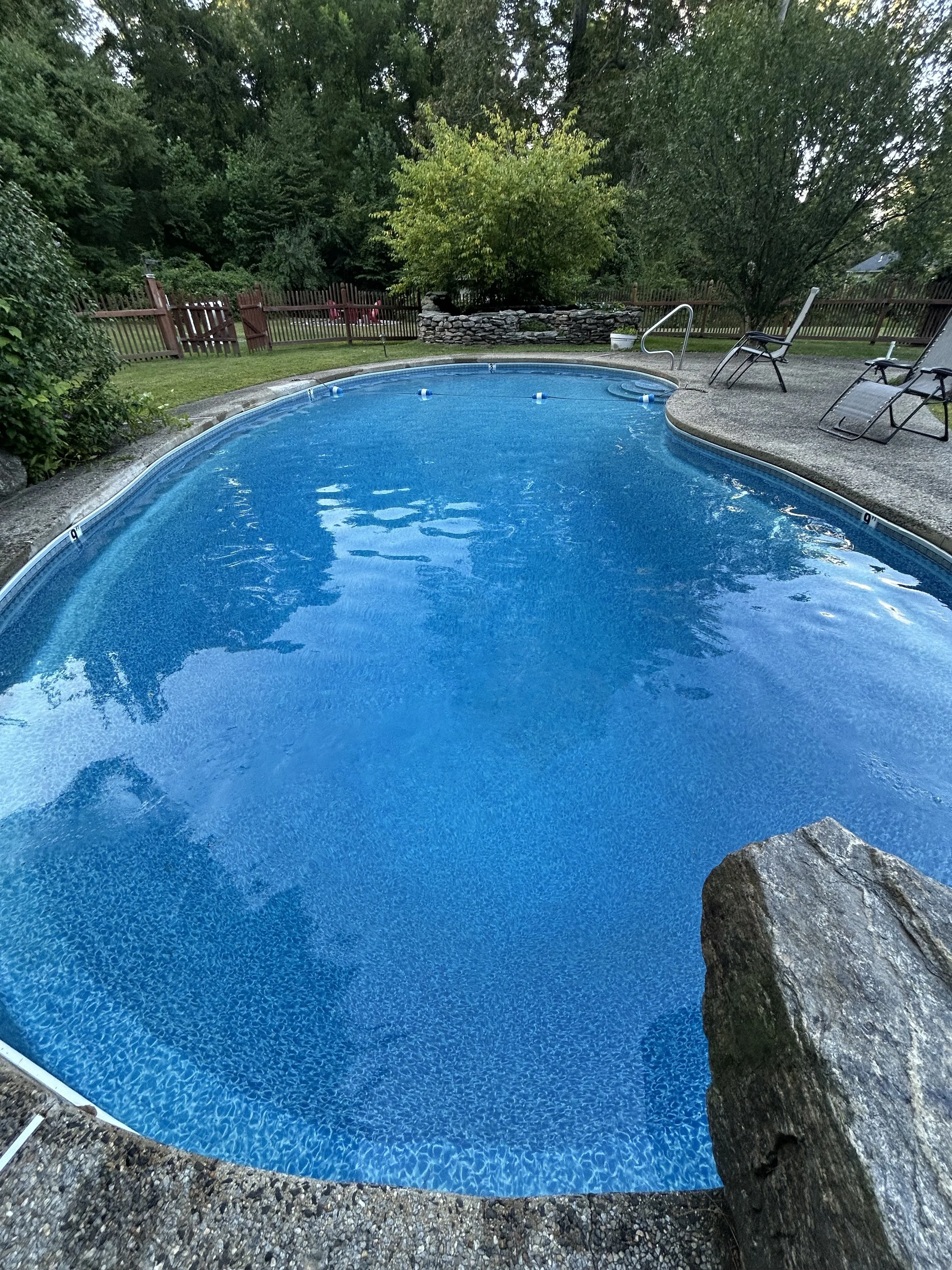 A backyard swimming pool surrounded by a concrete deck with a rock at the edge, lounge chairs, trees, and a wooden fence.