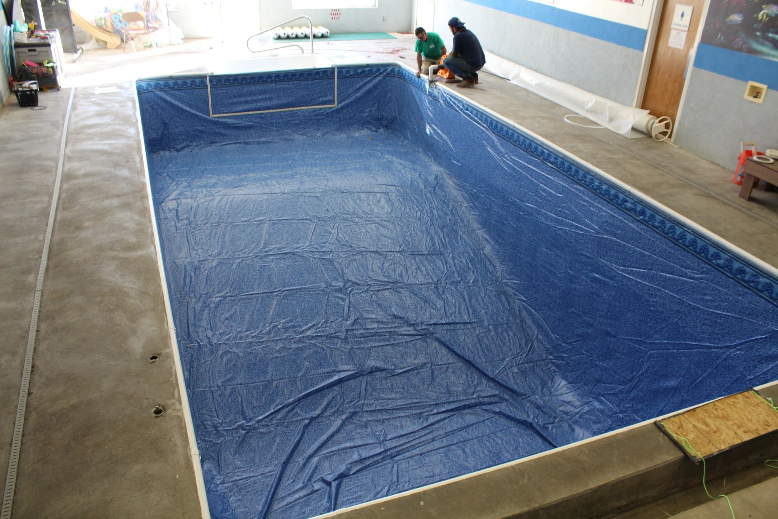 An indoor swimming pool under renovation, with a blue tarp lining the empty pool, workers in green and blue shirts working nearby, construction supplies, and tools around the pool area.