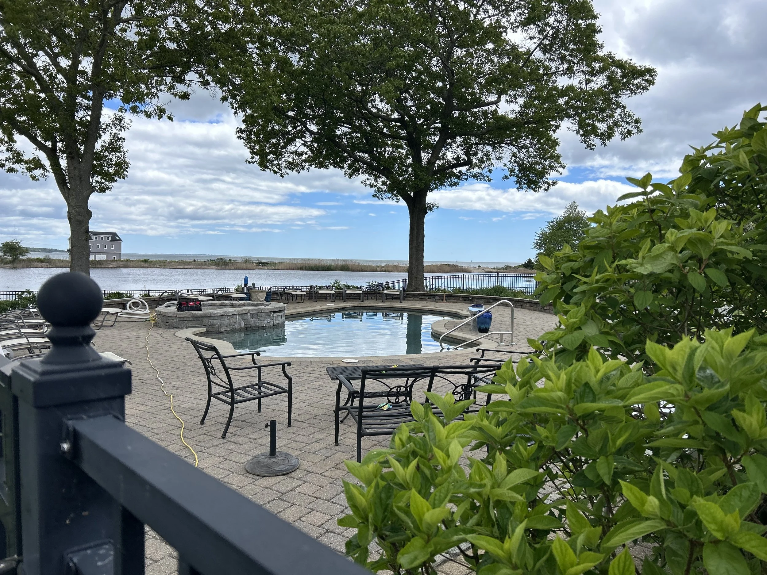 View of an outdoor pool area with a fire pit, surrounded by lounge chairs and trees, overlooking a body of water with a house in the distance under partly cloudy skies.