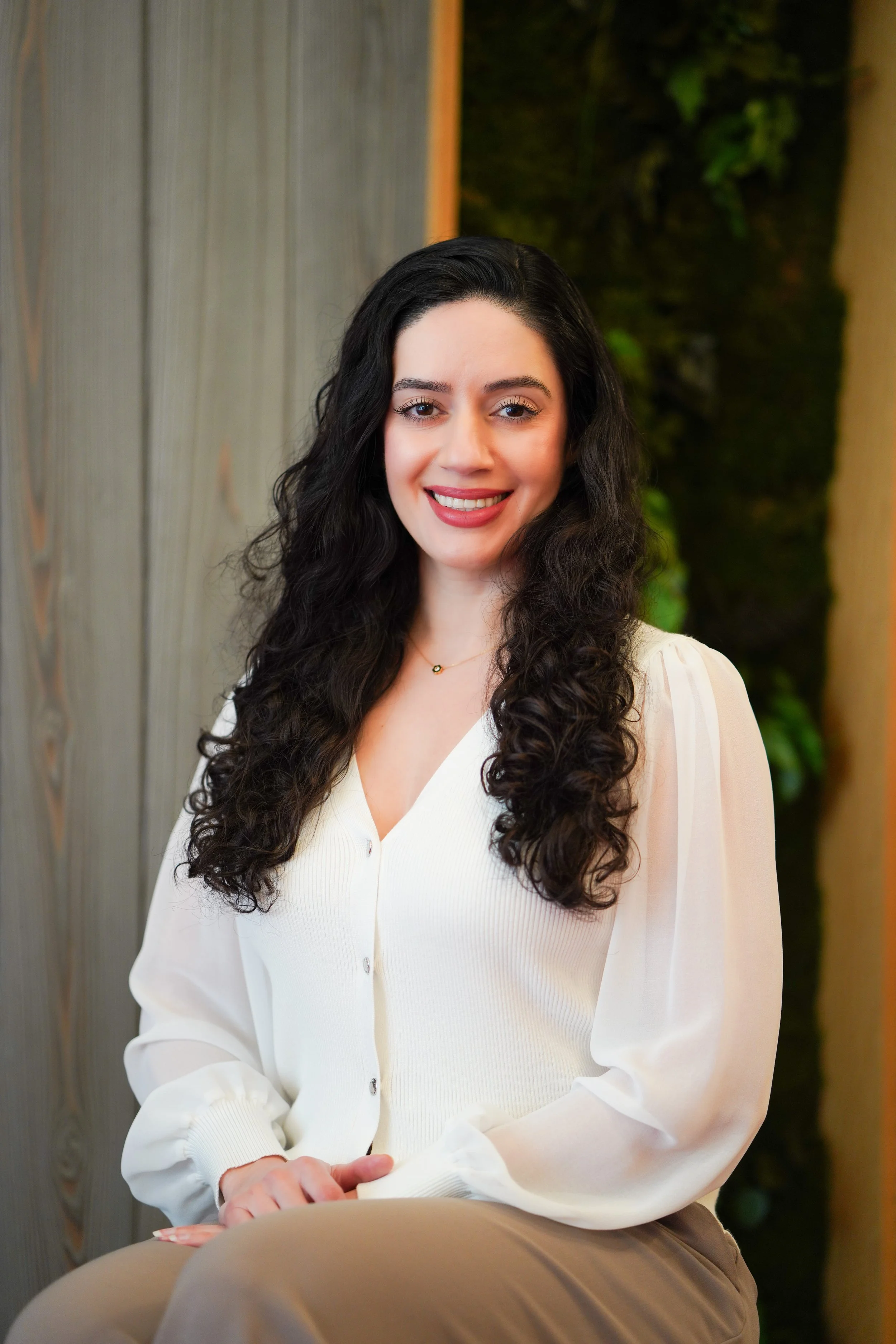 Smiling Setareh Kamali,  with long dark curly hair wearing a white blouse, seated indoors against a neutral background with greenery.