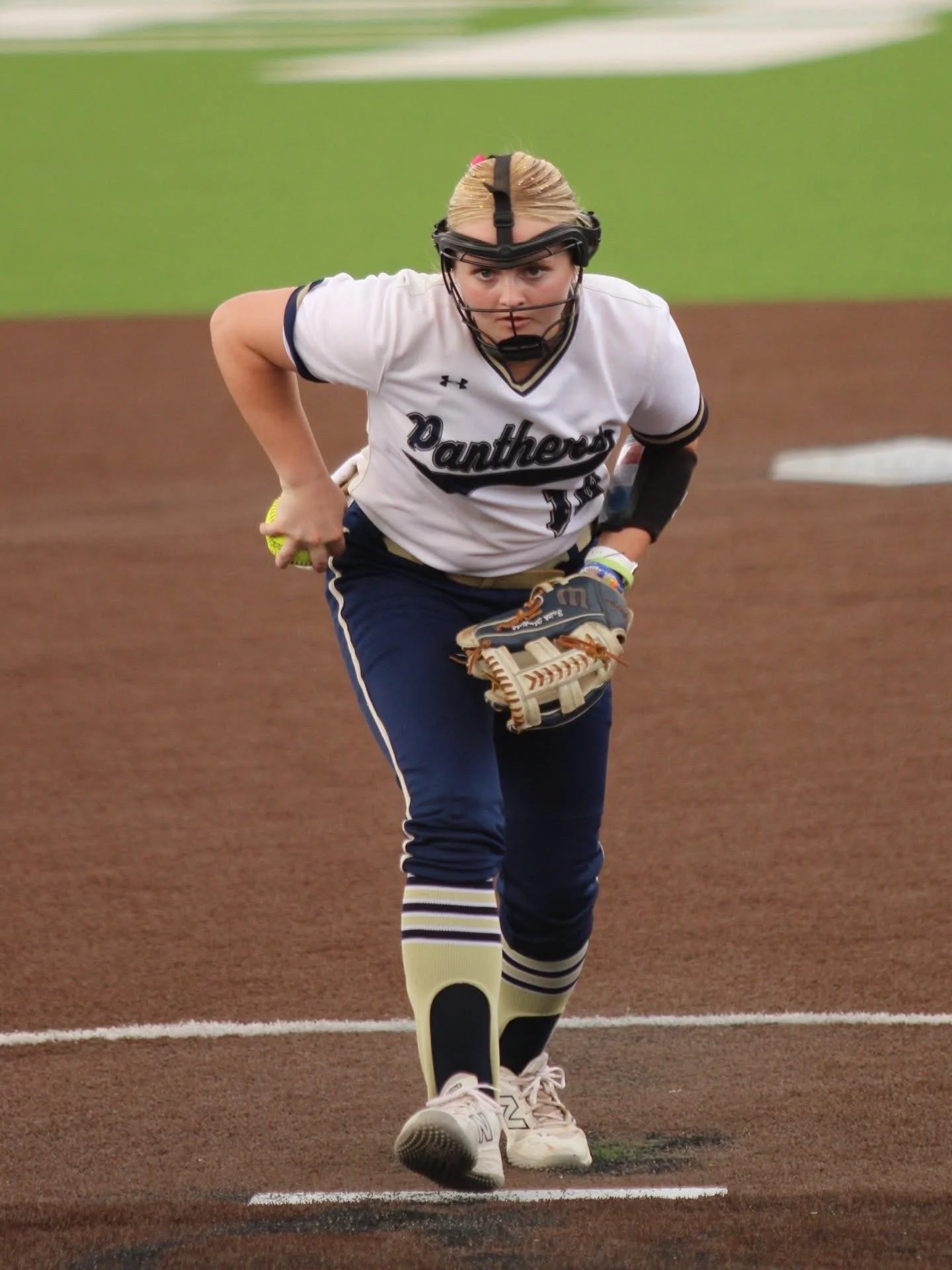 A female softball player wearing a white jersey with 'Panthers' written on it, navy blue pants, and striped knee-high socks, is running on a dirt field with a focused expression. She is holding a softball in her right hand and wearing a protective face mask and a baseball glove.