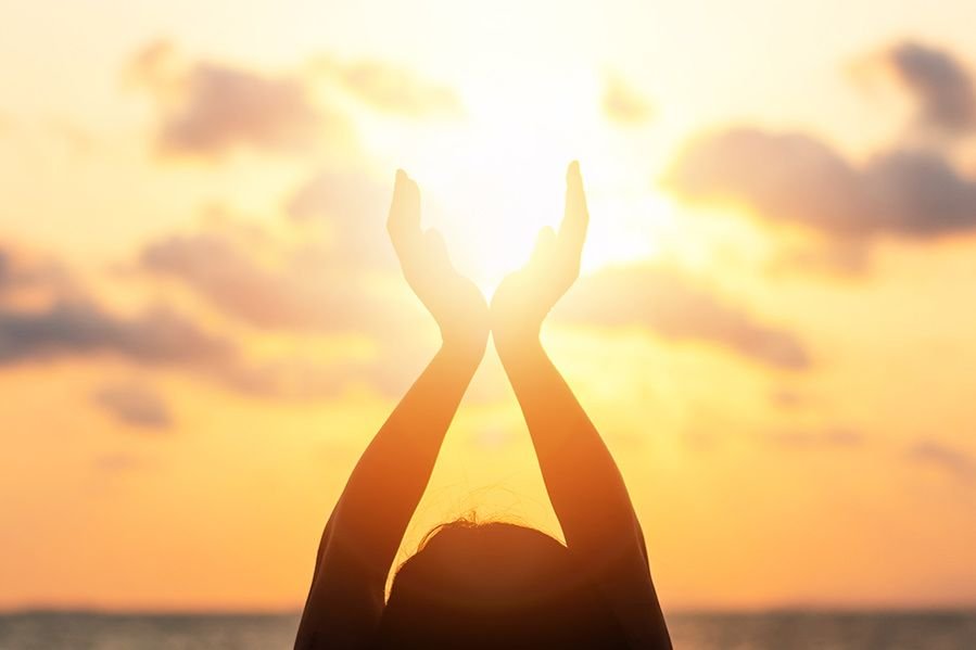Person holding hands up towards the sky with the sun shining between their hands during sunset or sunrise over the ocean.