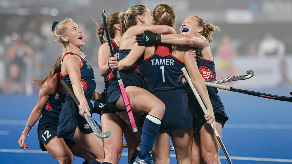 Female field hockey players celebrating and hugging on the field, wearing matching uniforms with one player's jersey reading 'Tamer 1'.