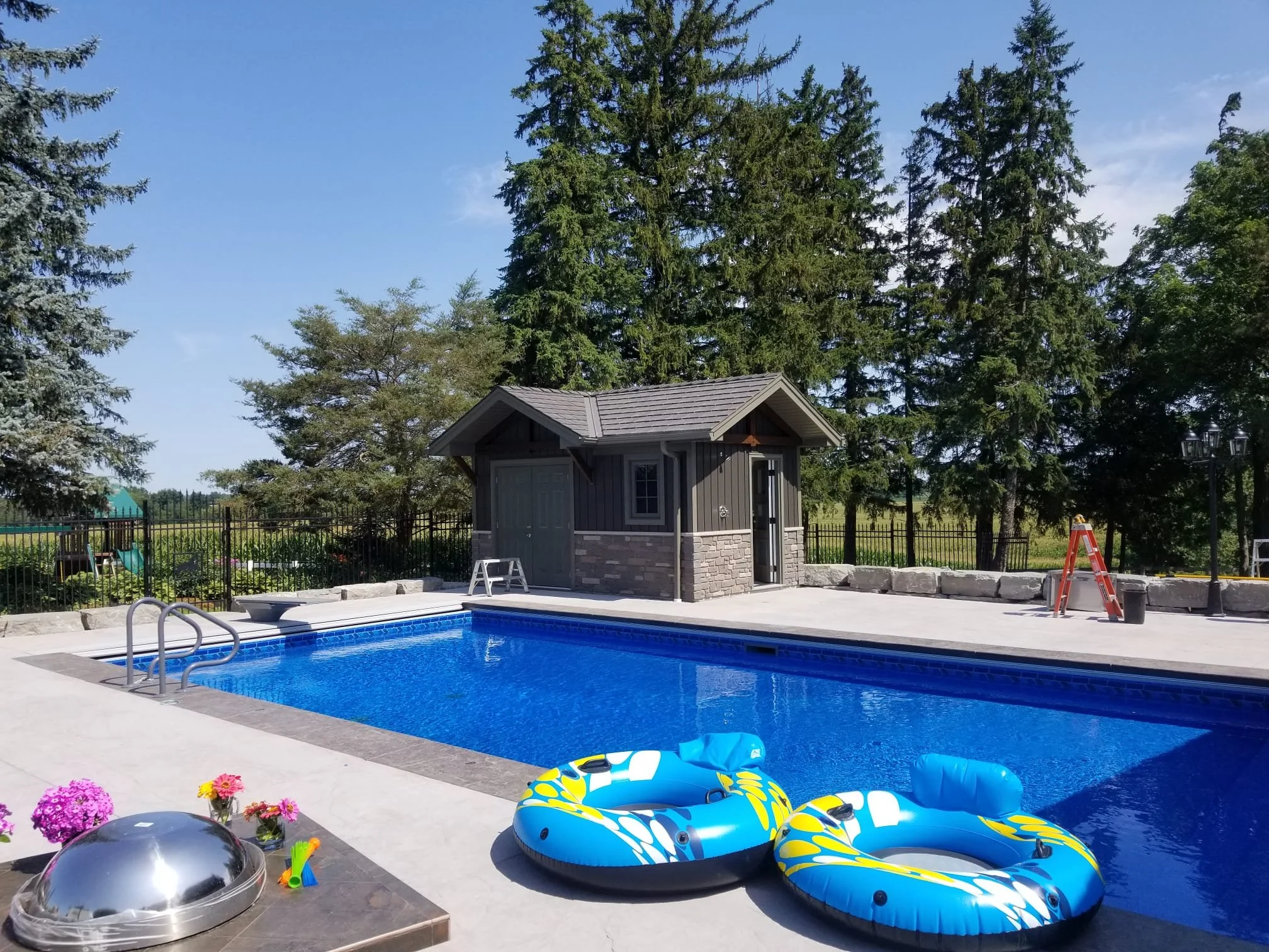 An outdoor backyard swimming pool with two blue inflatable floaties, surrounded by a concrete deck, with a small wooden pool house, trees, flowers, and a fence in the background.