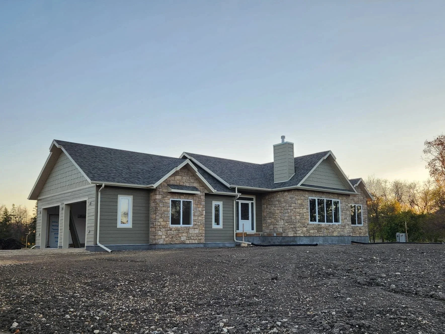 Newly constructed single-family house with stone and siding exterior, large windows, and a gravel driveway during sunset.