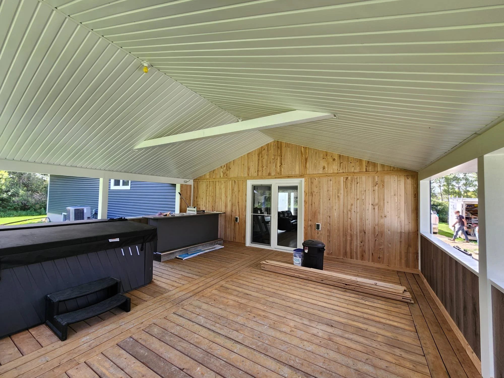 An outdoor covered porch with a wooden deck, a hot tub, and a sliding glass door leading into a house with wooden wall paneling and white window frames. Construction materials and a black trash bin are visible, with some people and a truck outside.