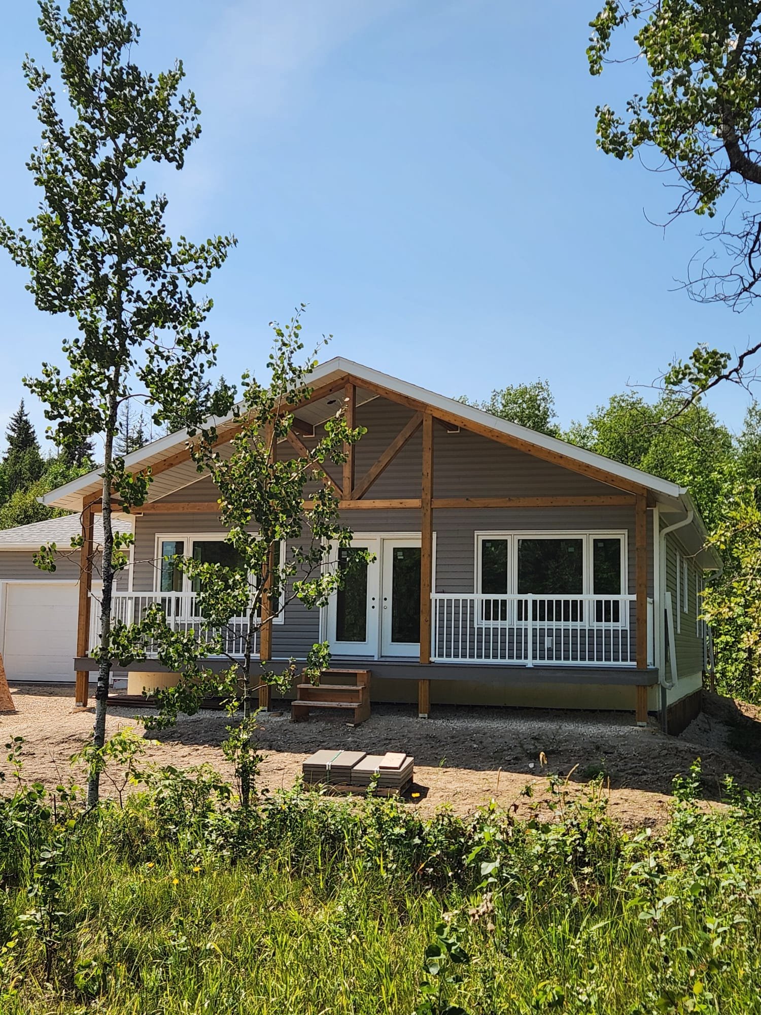 Front view of a house under construction with a covered porch, white doors, gray siding, and a partially built wooden structure for the roof, surrounded by trees and early landscaping.