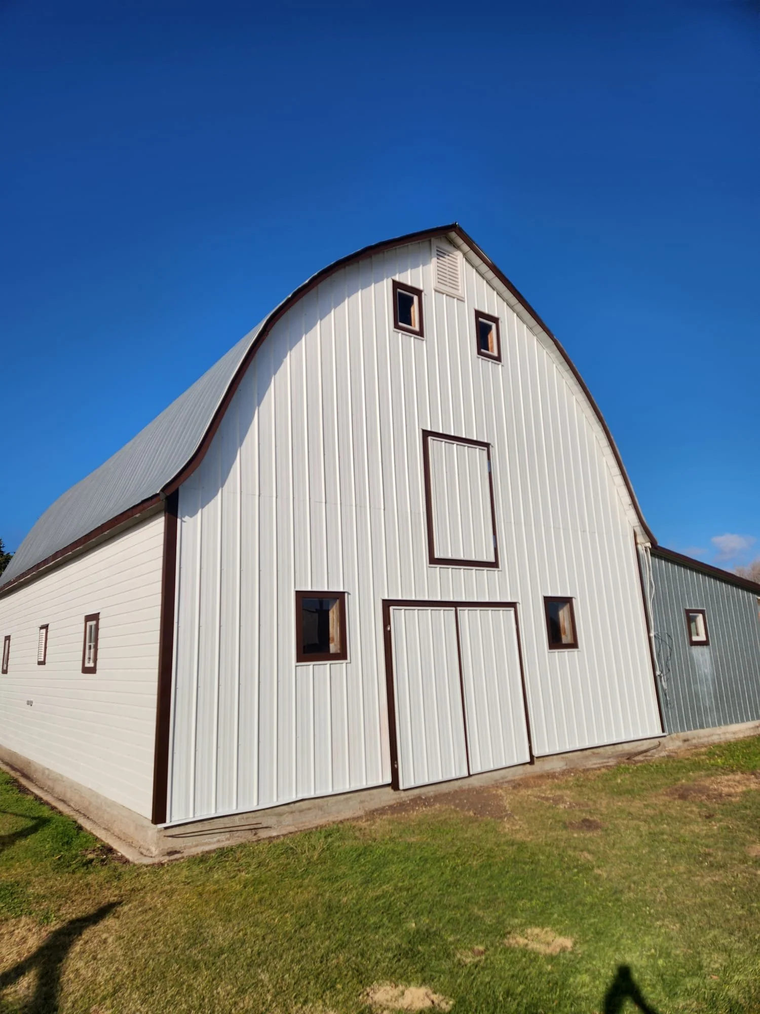 A large white barn with a curved roof, small windows, and a central door, set against a clear blue sky and grassy ground.