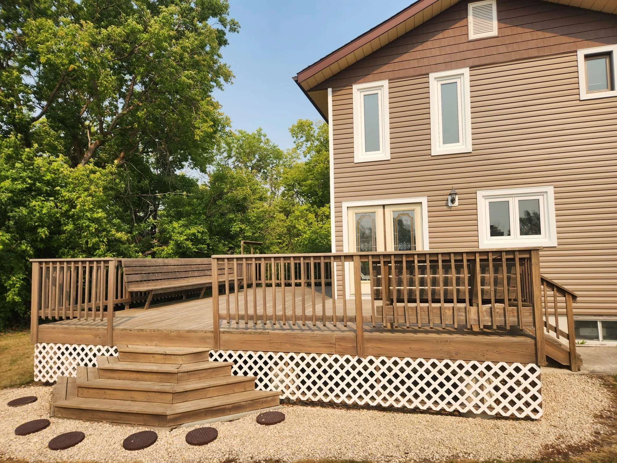 A wooden deck with stairs leading up to it, attached to a beige house with window and glass door, surrounded by green trees and a gravel yard.