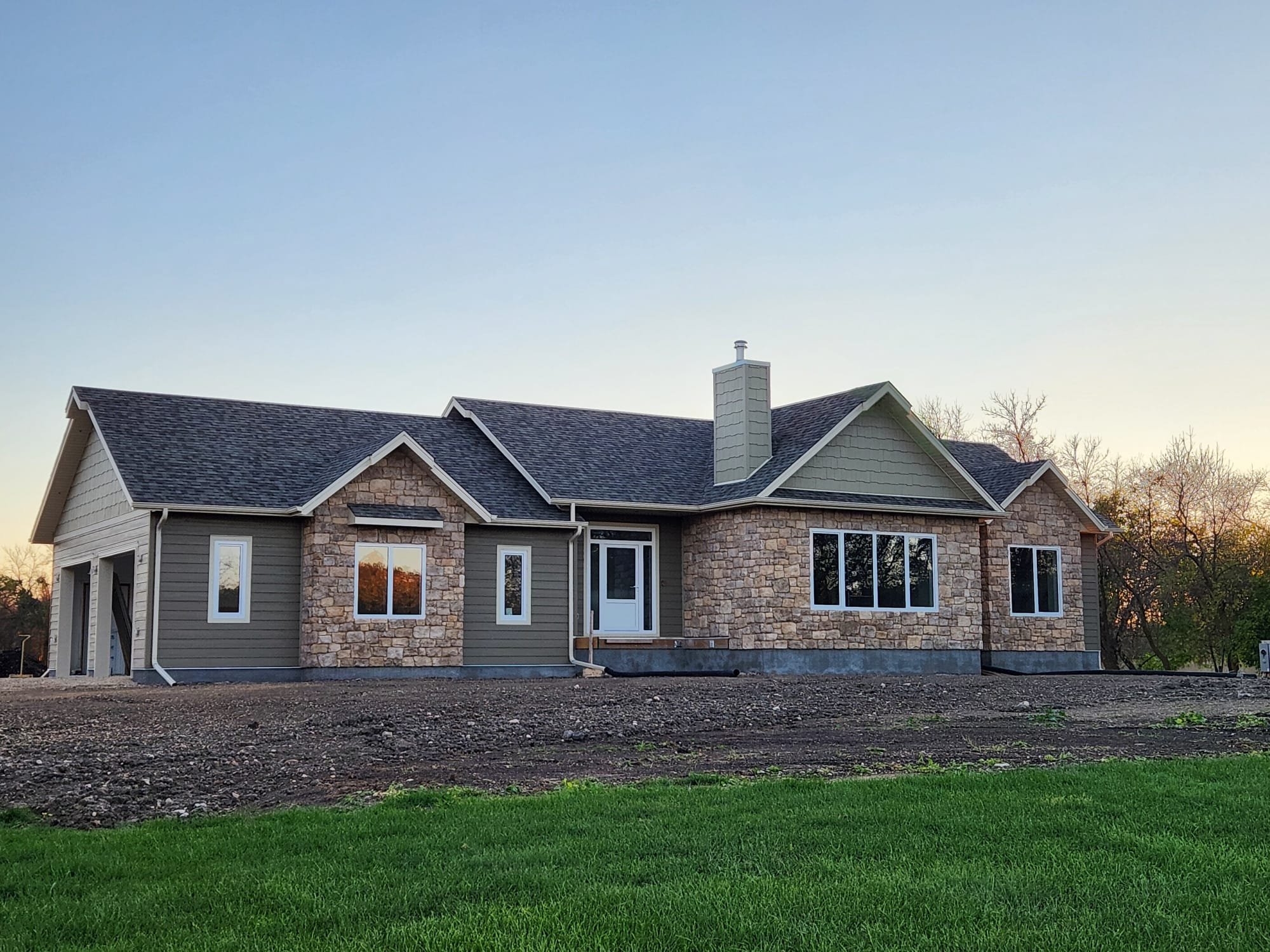 New house under construction with stone and siding exterior, multiple windows, and a chimney in a suburban area with trees in the background at sunset.
