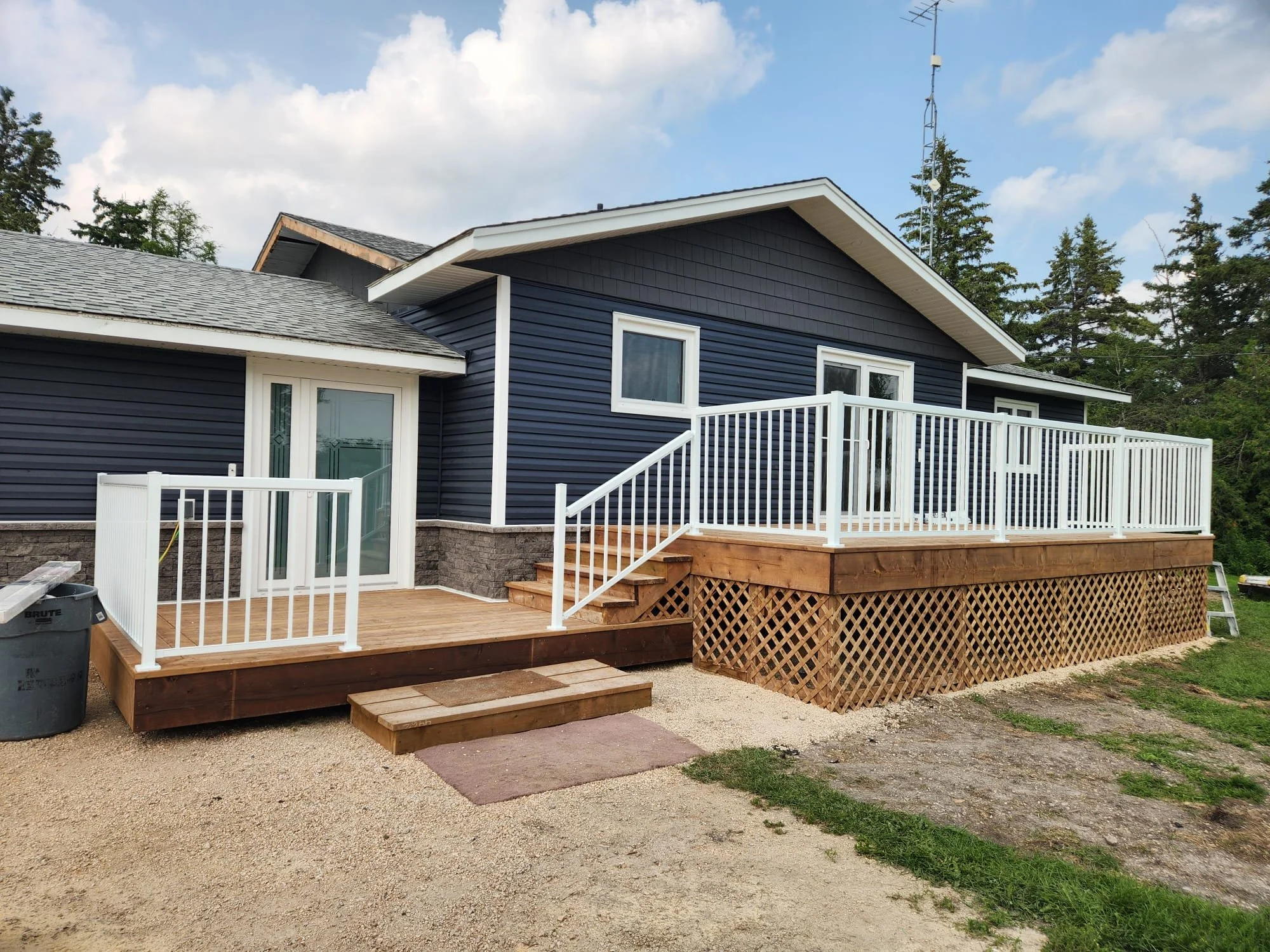 Newly built wooden deck with white railing attached to a blue house with sliding glass door, surrounded by a gravel yard and trees.