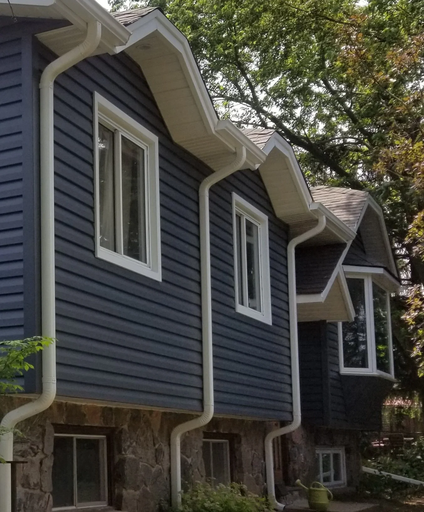 Photo of the side of a house with navy blue siding, white trimmed windows, and white gutters, surrounded by trees.