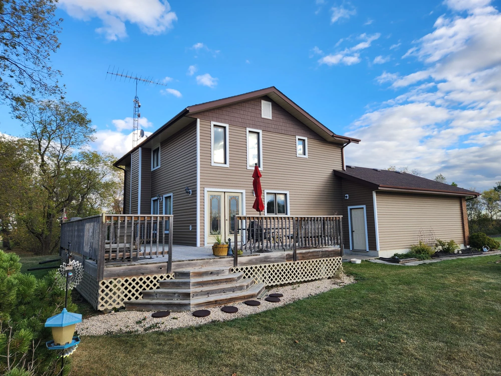 A two-story house with beige siding and a deck in back with steps leading down to a grassy yard. The deck has a red patio umbrella and outdoor furniture. There are trees and a blue sky with clouds in the background.