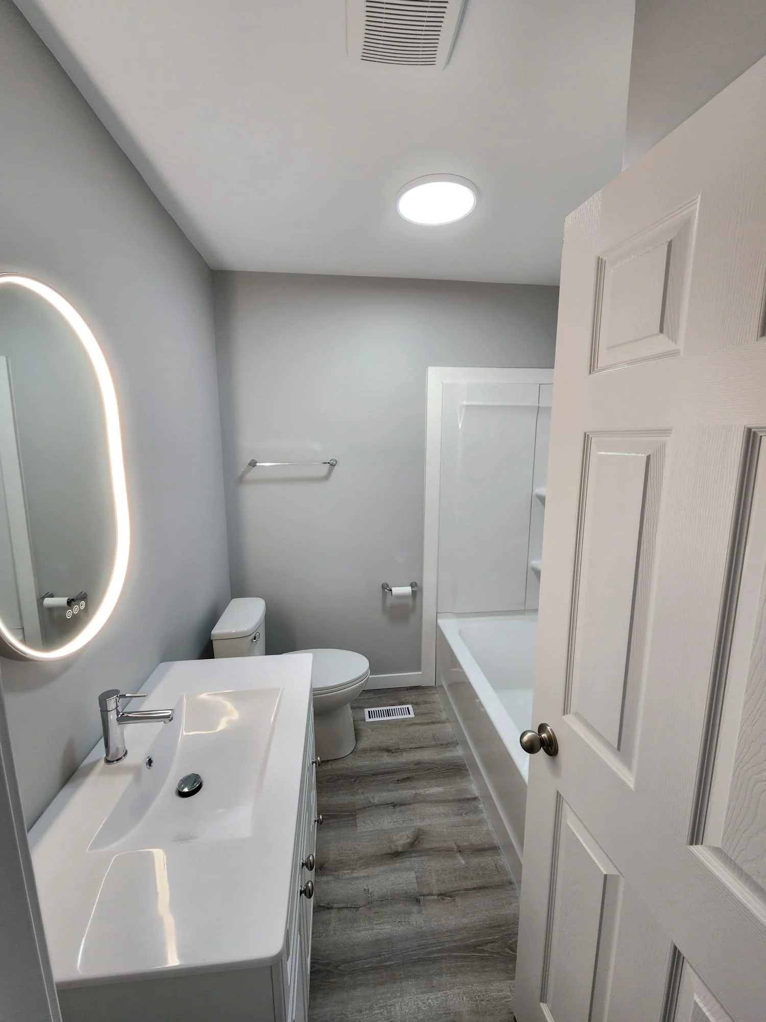 A modern bathroom with a white vanity, a mirror with LED lighting, a toilet, a bathtub with a shower curtain, and wood-look flooring.