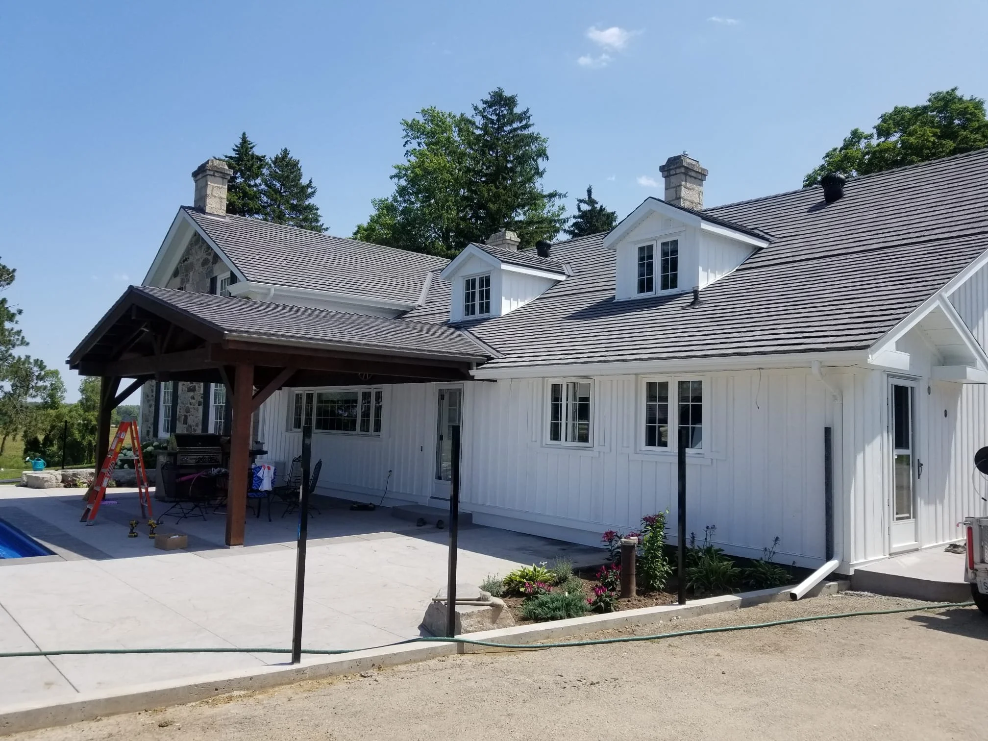 Newly renovated white house with dark gray roof, covered outdoor patio with dining table and chairs, surrounded by green trees and plants, under a clear blue sky.