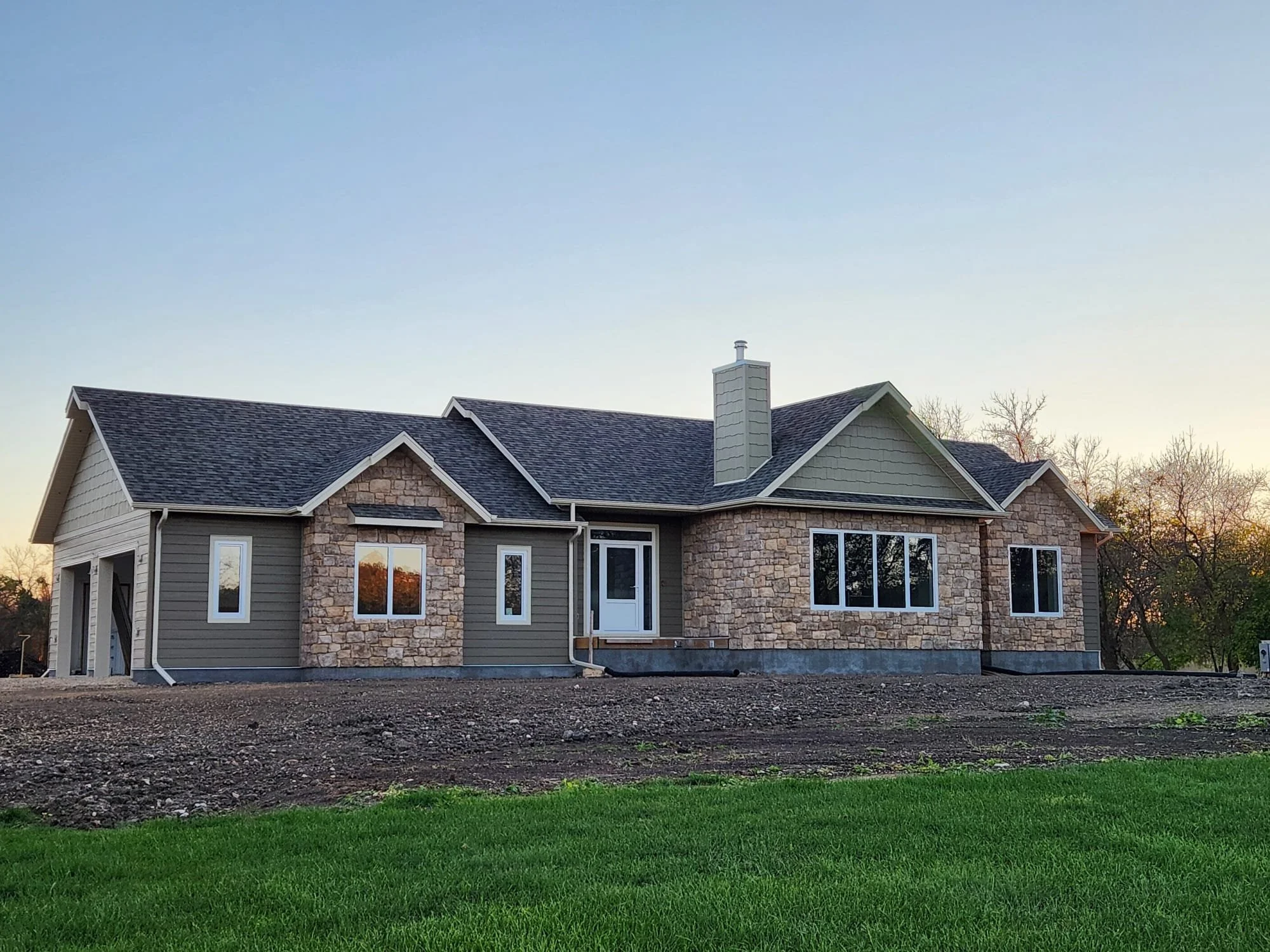 New house under construction with a stone and siding exterior, multiple windows, and a chimney, set against a sunset sky with trees in the background.