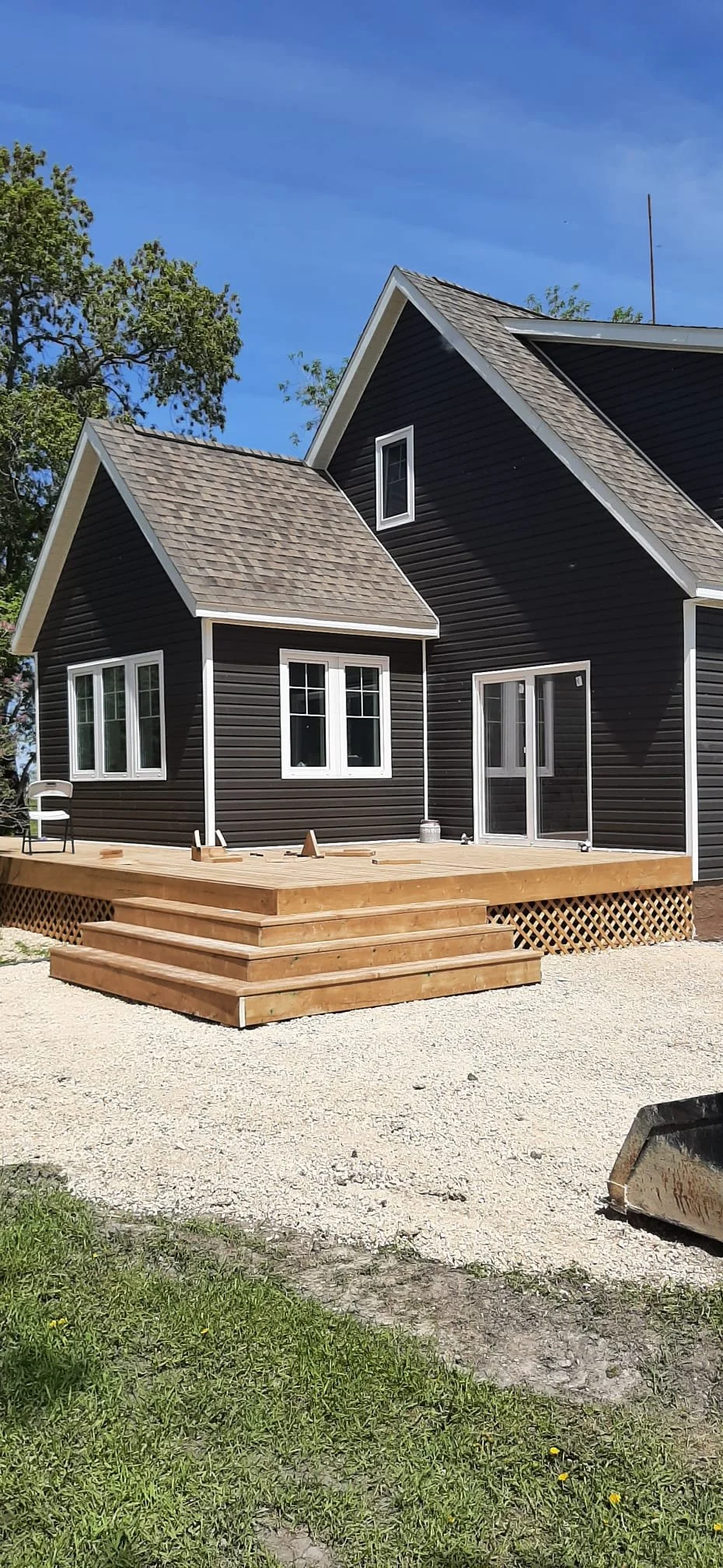 A new black house with a wooden deck, stairs, and a gravel yard, set against a background of green trees and a blue sky.