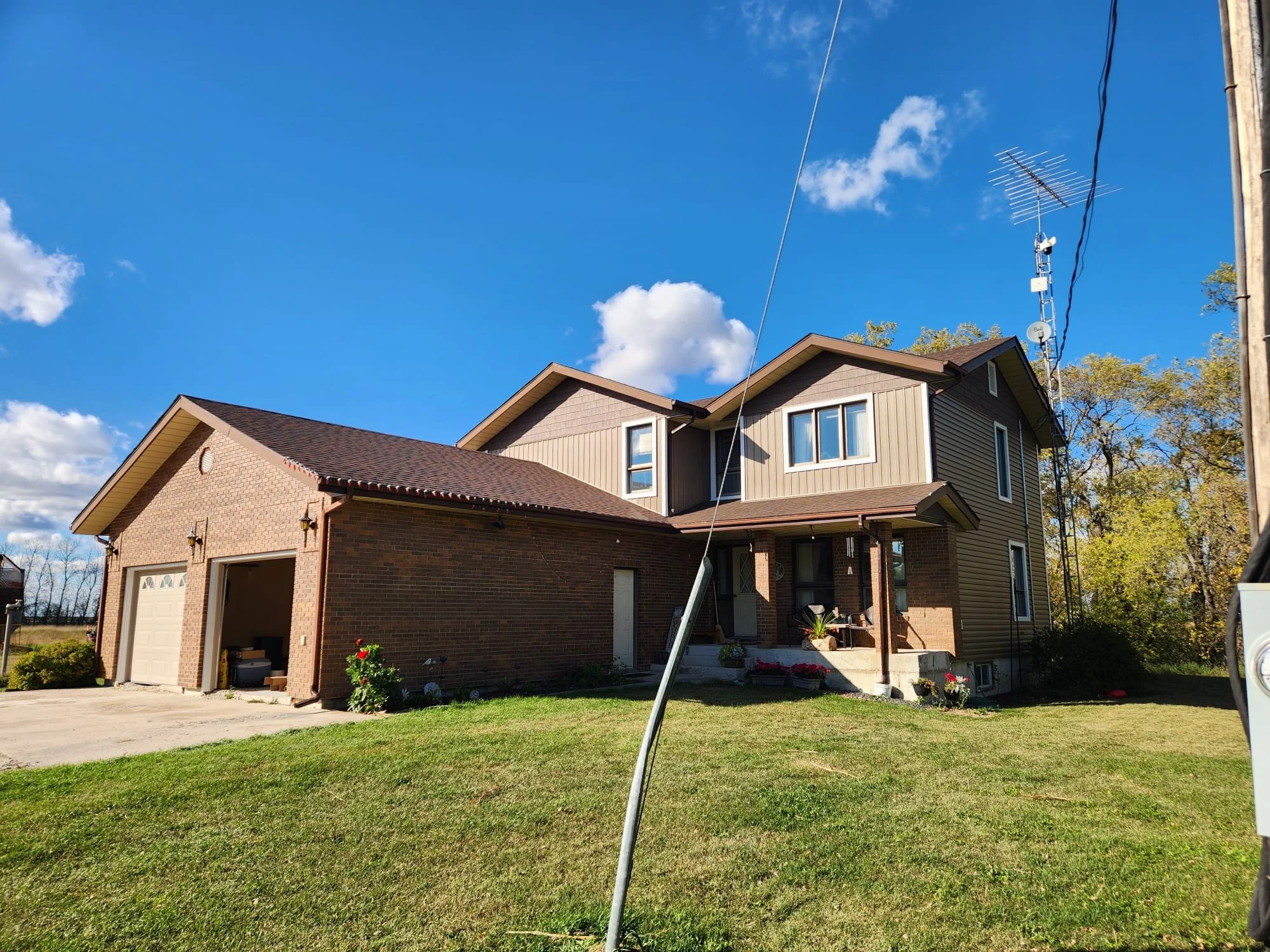 A two-story house with a brick garage, tan siding, and a small front porch with potted plants. A lawn and driveway are visible in the foreground, and there are trees and a blue sky with white clouds in the background. An antenna is mounted on a pole 