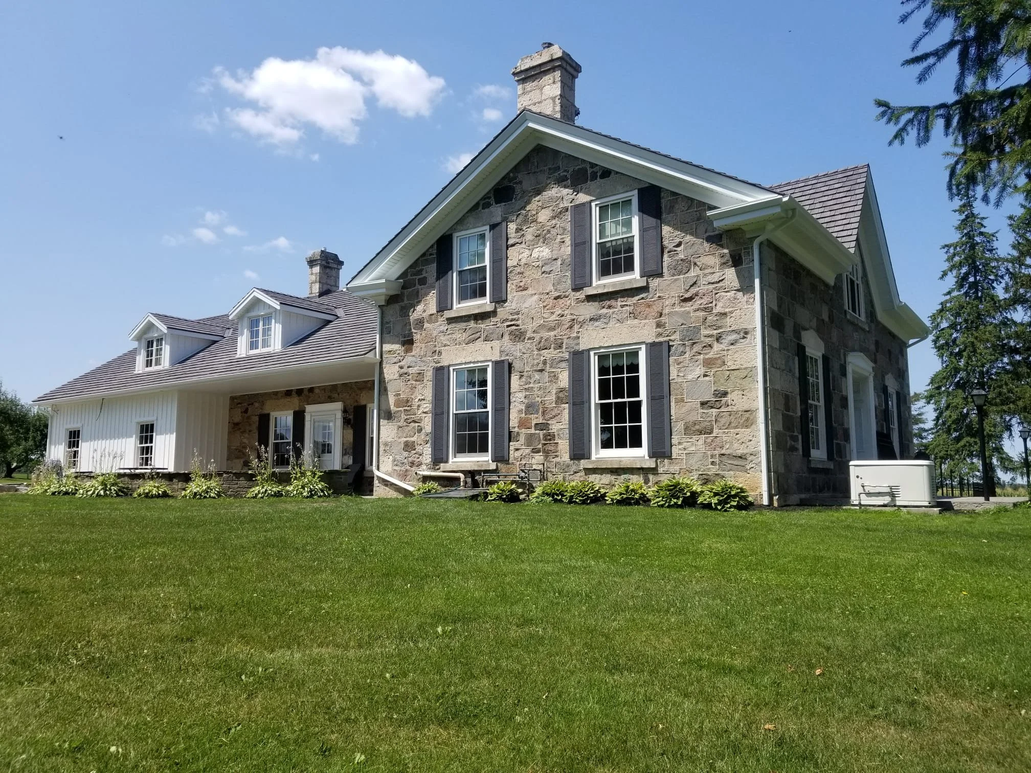 A two-story house with a stone exterior, black shutters, and a gray shingle roof, set on a well-manicured lawn with a blue sky in the background.