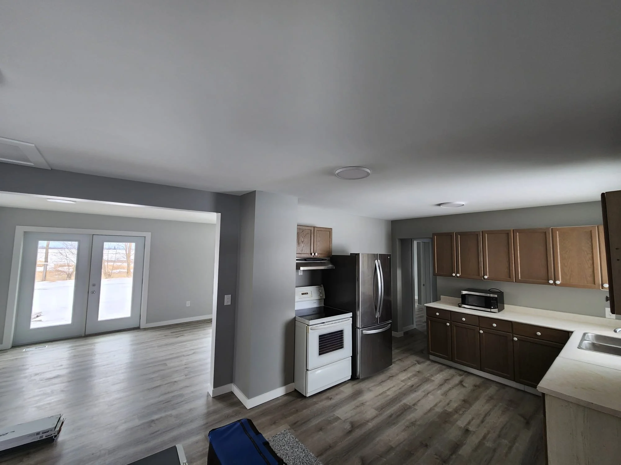Open-concept living area with kitchen featuring wooden cabinets, stainless steel refrigerator, white stove, microwave, and a view of double glass doors leading outside, with white walls and gray ceiling.
