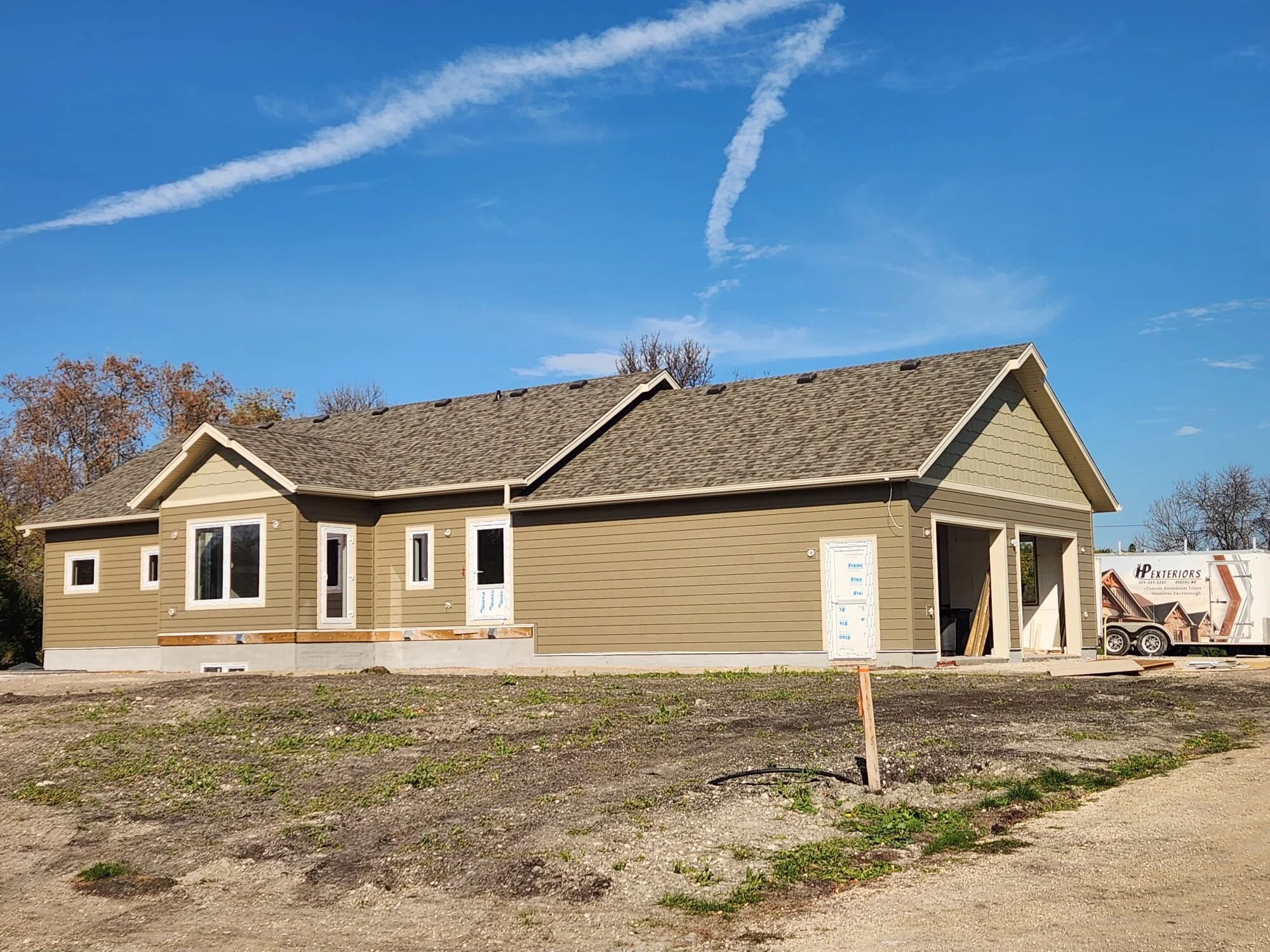 A house under construction with beige siding and a brown shingled roof, located in a construction site with dirt and sparse grass, and a trailer in the background.