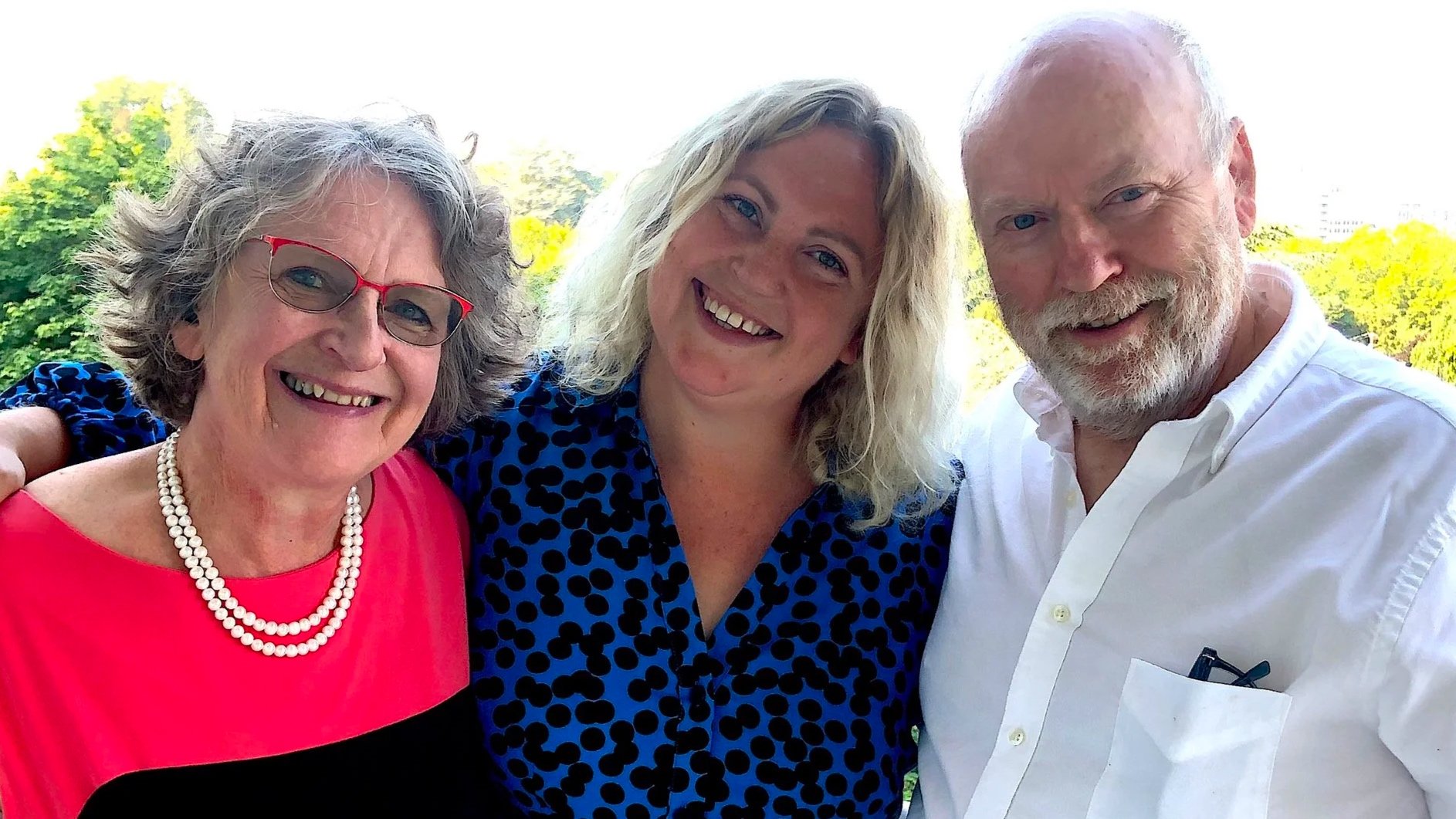 Three smiling people, two women and one man, posing outdoors with trees and blue sky in the background. This is Frani, Martha, and Mike Grover.