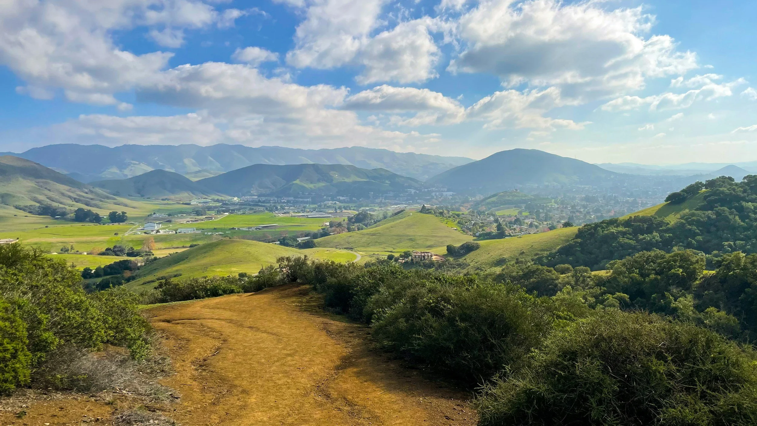 View from a hillside dirt trail overlooking rolling green hills, farmland, and a town in a valley, with mountains in the distance under a blue sky with scattered clouds.