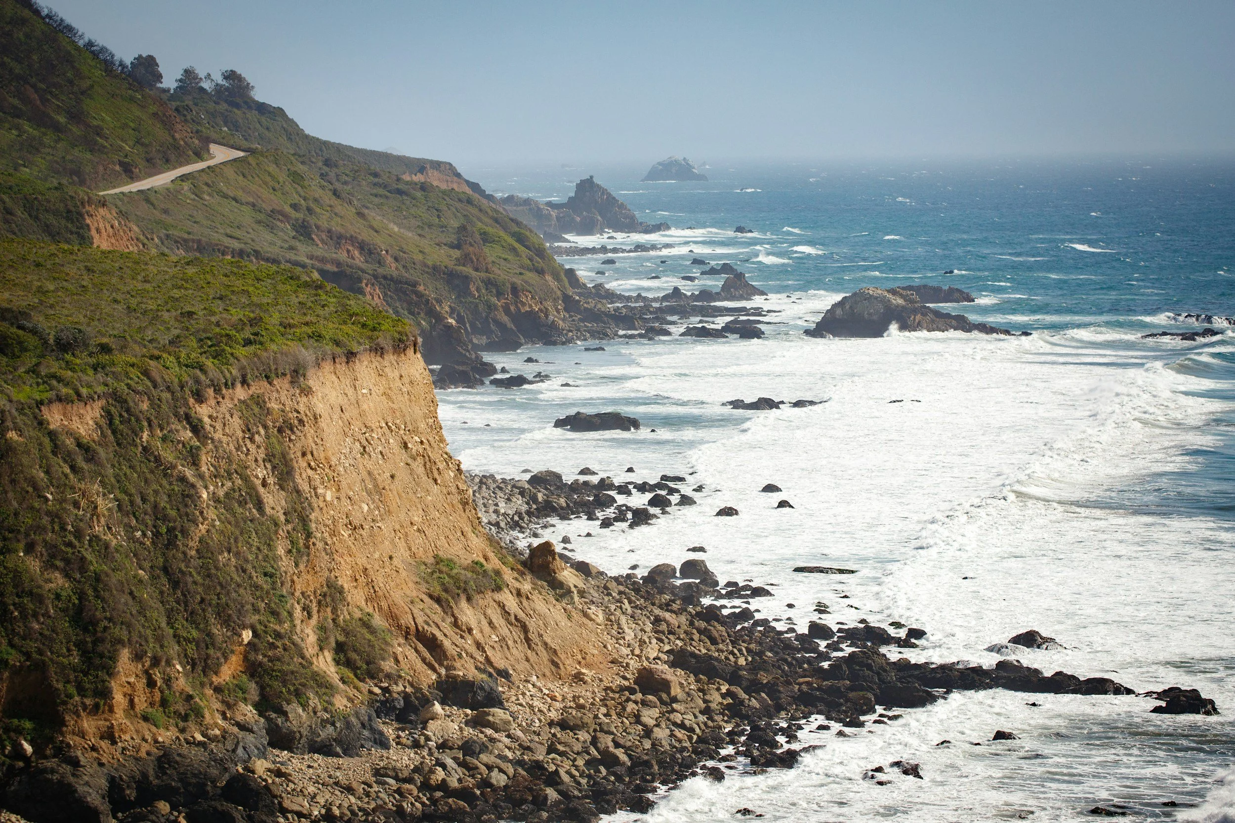 View of scenic central coast cliffs and a rocky shoreline with waves breaking along the coast, with the ocean and distant rock formations under a hazy sky.