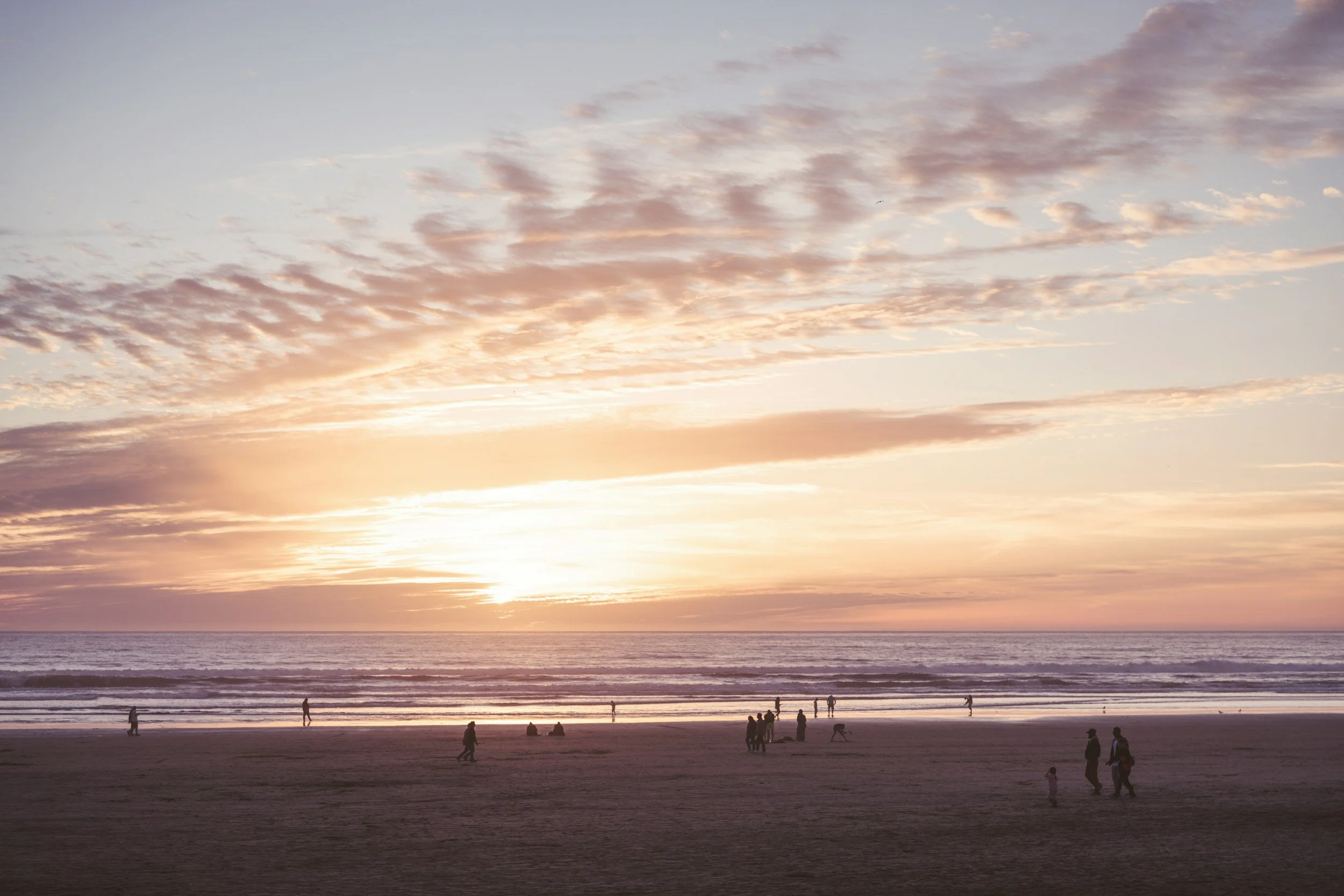 People walking and gathering on a beach at sunset, with soft waves and pastel clouds over the ocean.