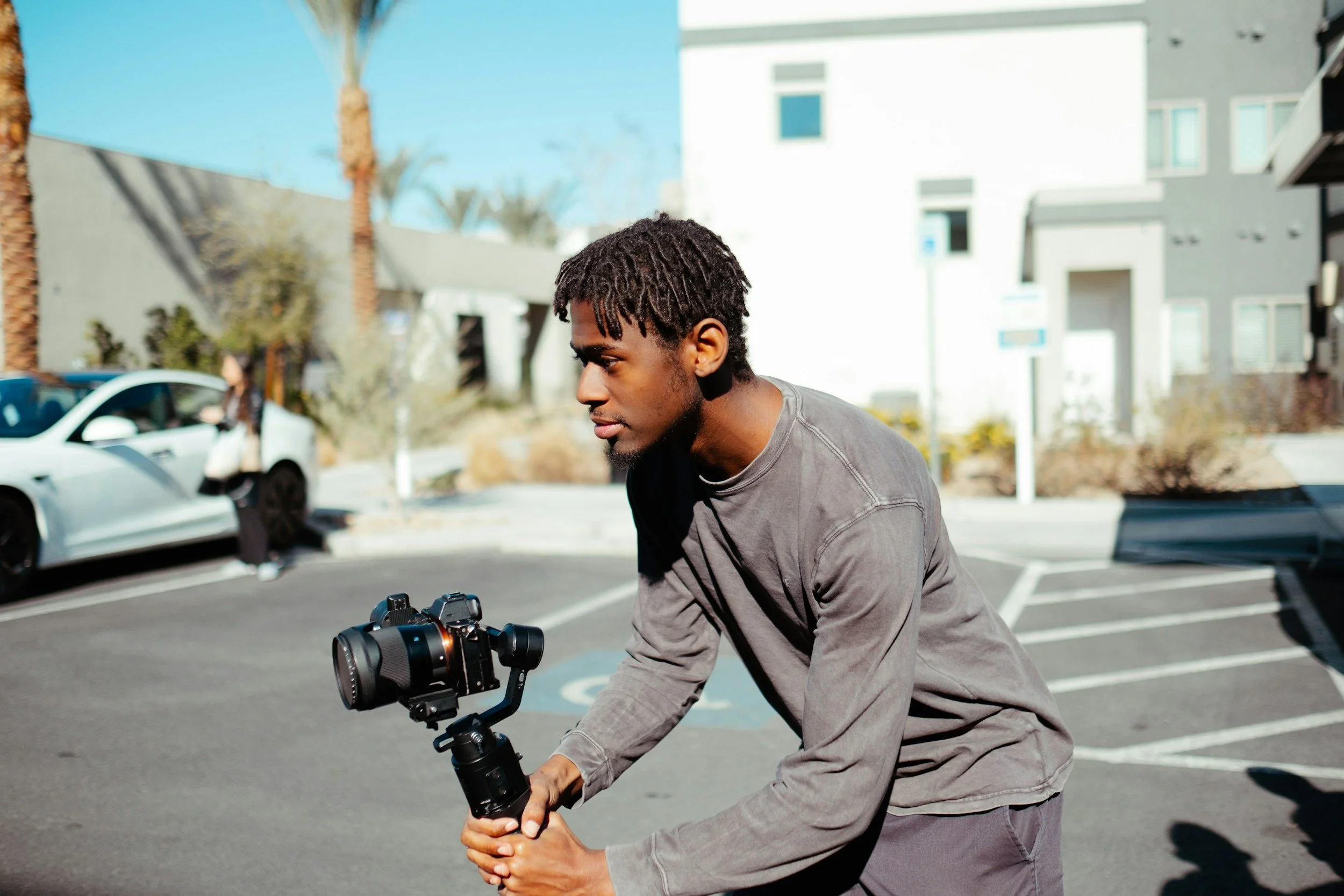 Young man adjusting a professional camera mounted on a gimbal in an outdoor parking lot with modern buildings and palm trees in the background.