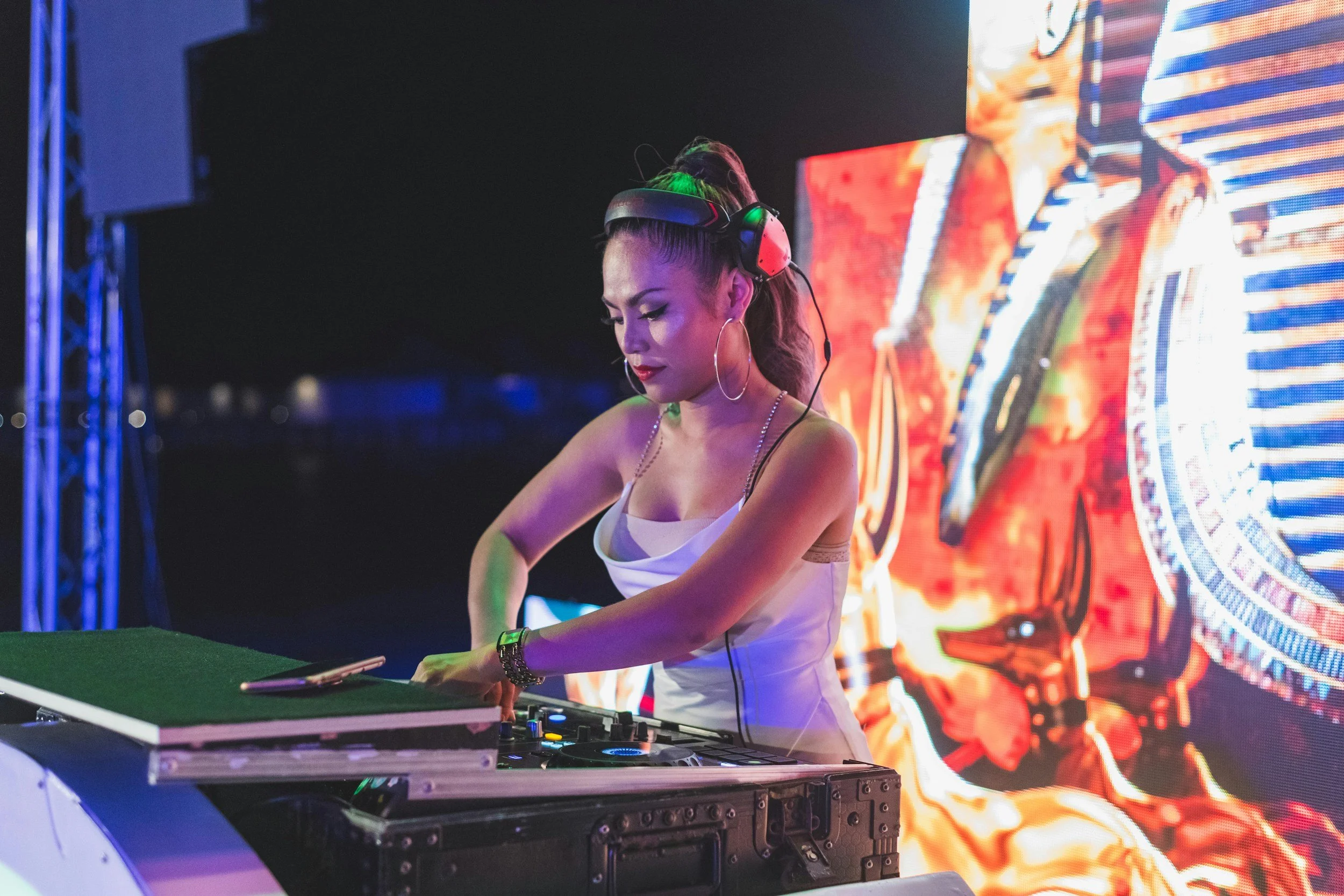 A female DJ with long hair tied in a high ponytail, wearing large hoop earrings and headphones, is working at her DJ booth during a night event with colorful LED screens in the background.