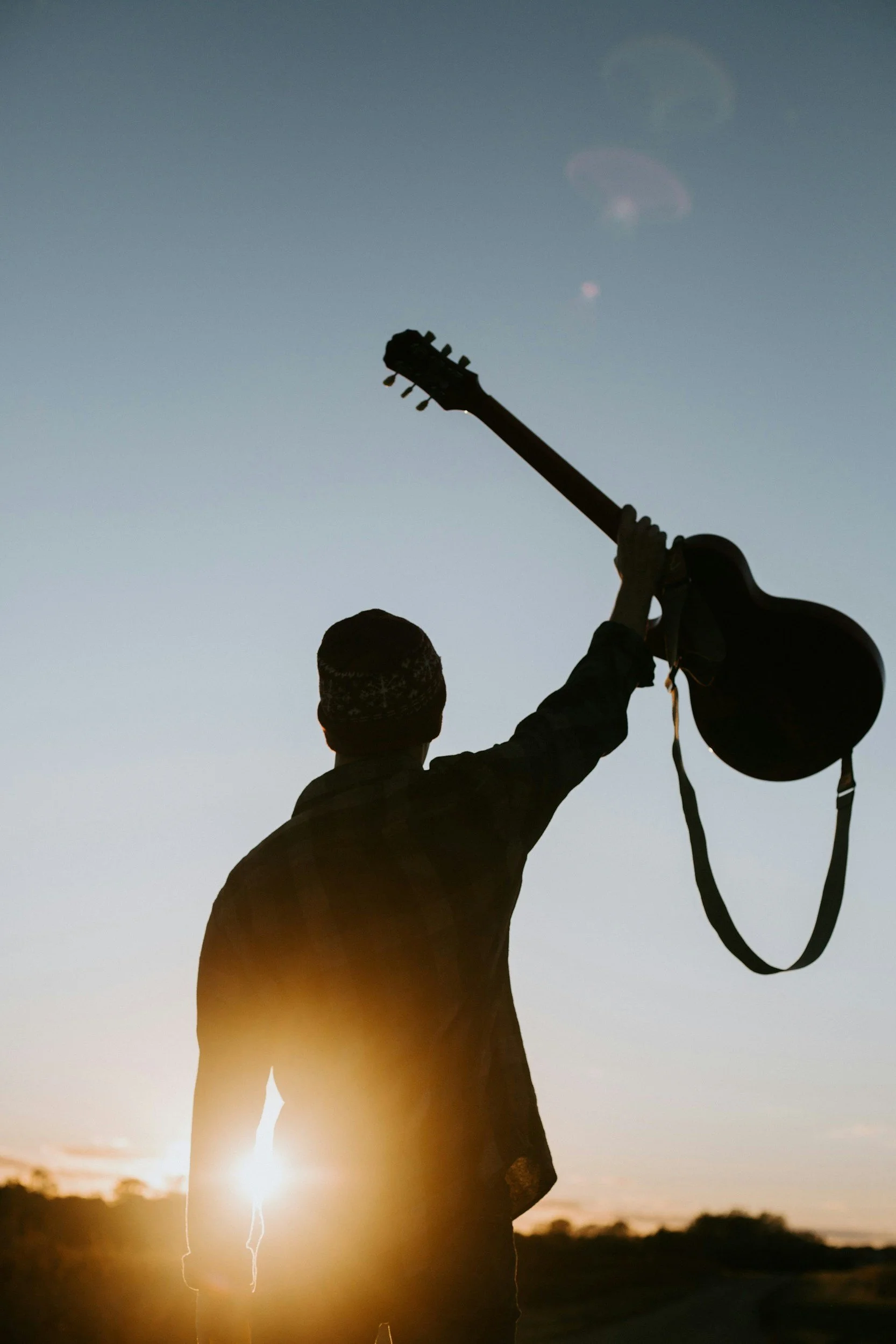 Person holding a guitar and a helmet outside during sunset.