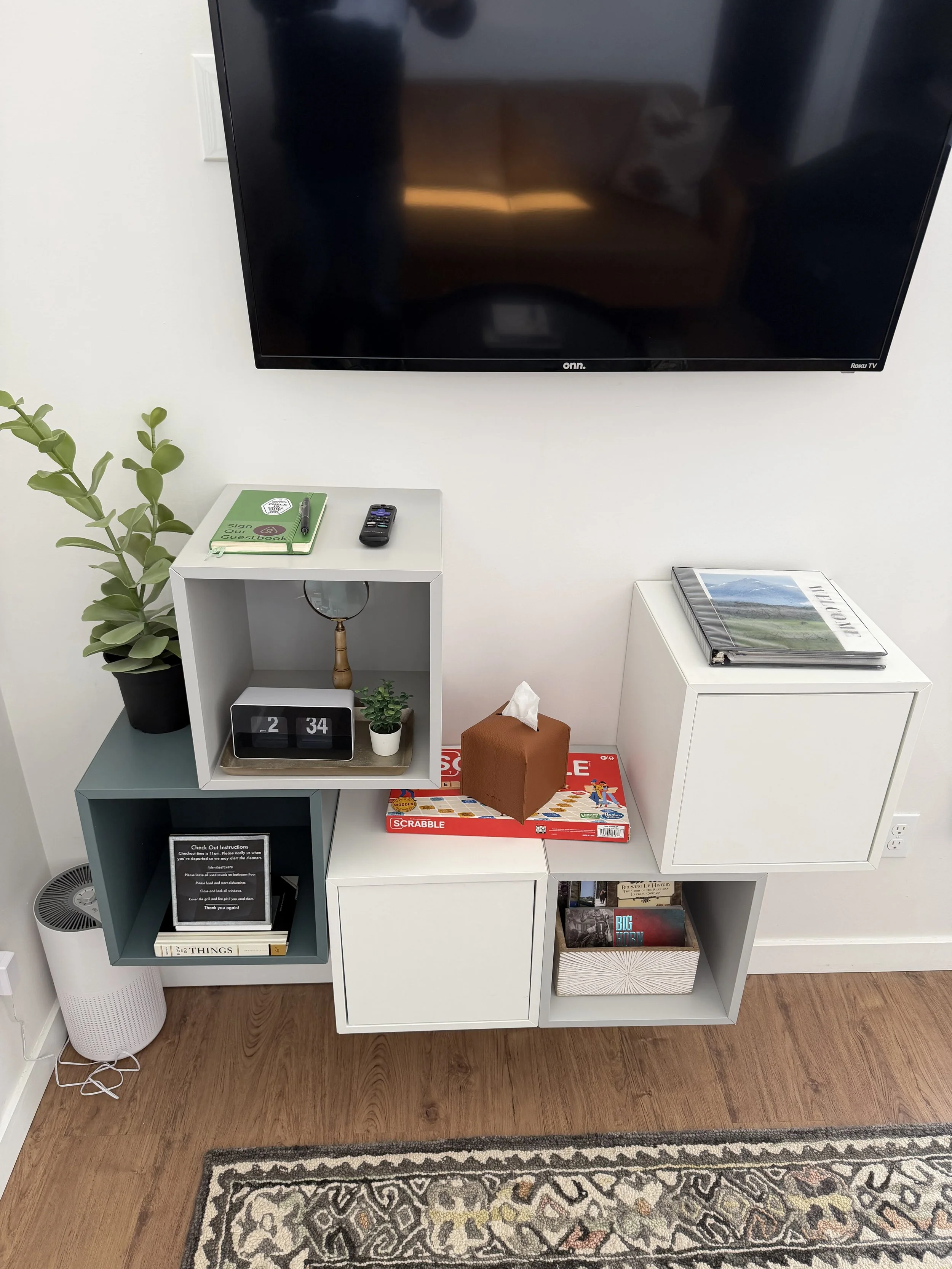 Modern entertainment center with a large flat-screen TV mounted on the wall above it, decorated with books, a tissue box, a clock, a plant, a note, and a remote control, with a patterned rug on a wooden floor.