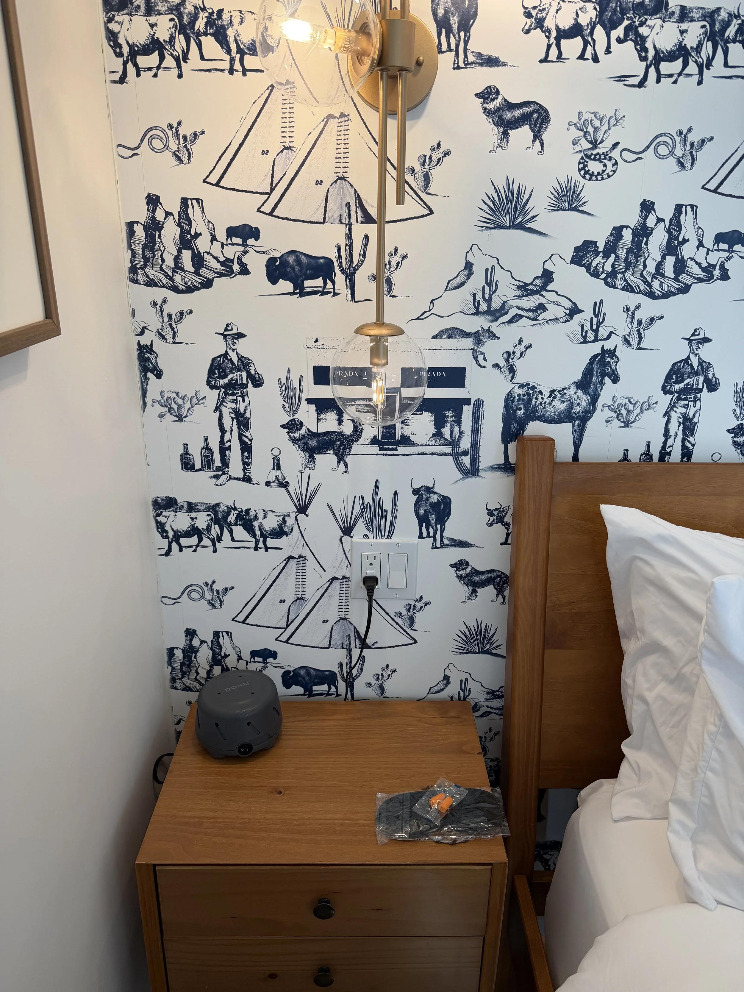 A bedroom nightstand with a gray alarm clock and a small plastic bag with an orange item on it. Above, a wall lamp with a round glass shade is mounted on a wallpaper featuring a blue and white Western desert scene with cowboys, horses, cacti, mountains, tents, and other desert elements.