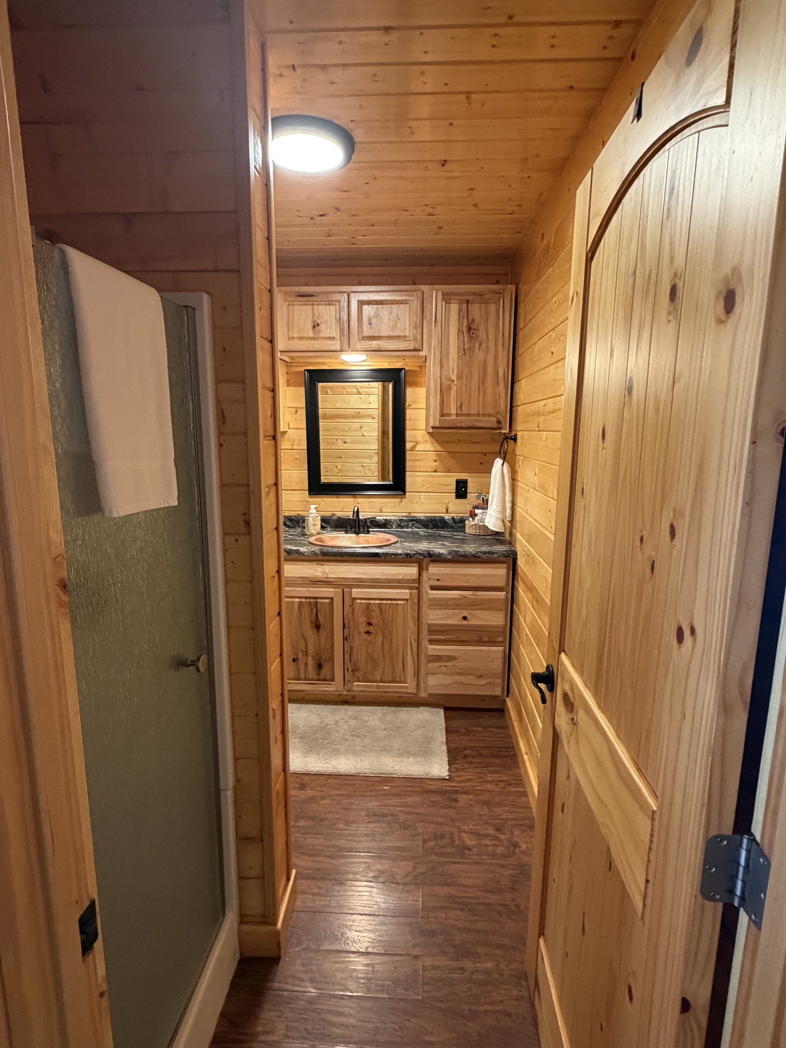 View of a rustic bathroom with wooden walls and ceiling, a black-framed mirror above a wooden vanity with a black faucet, a soap dispenser, and a small rug on a dark wood floor.