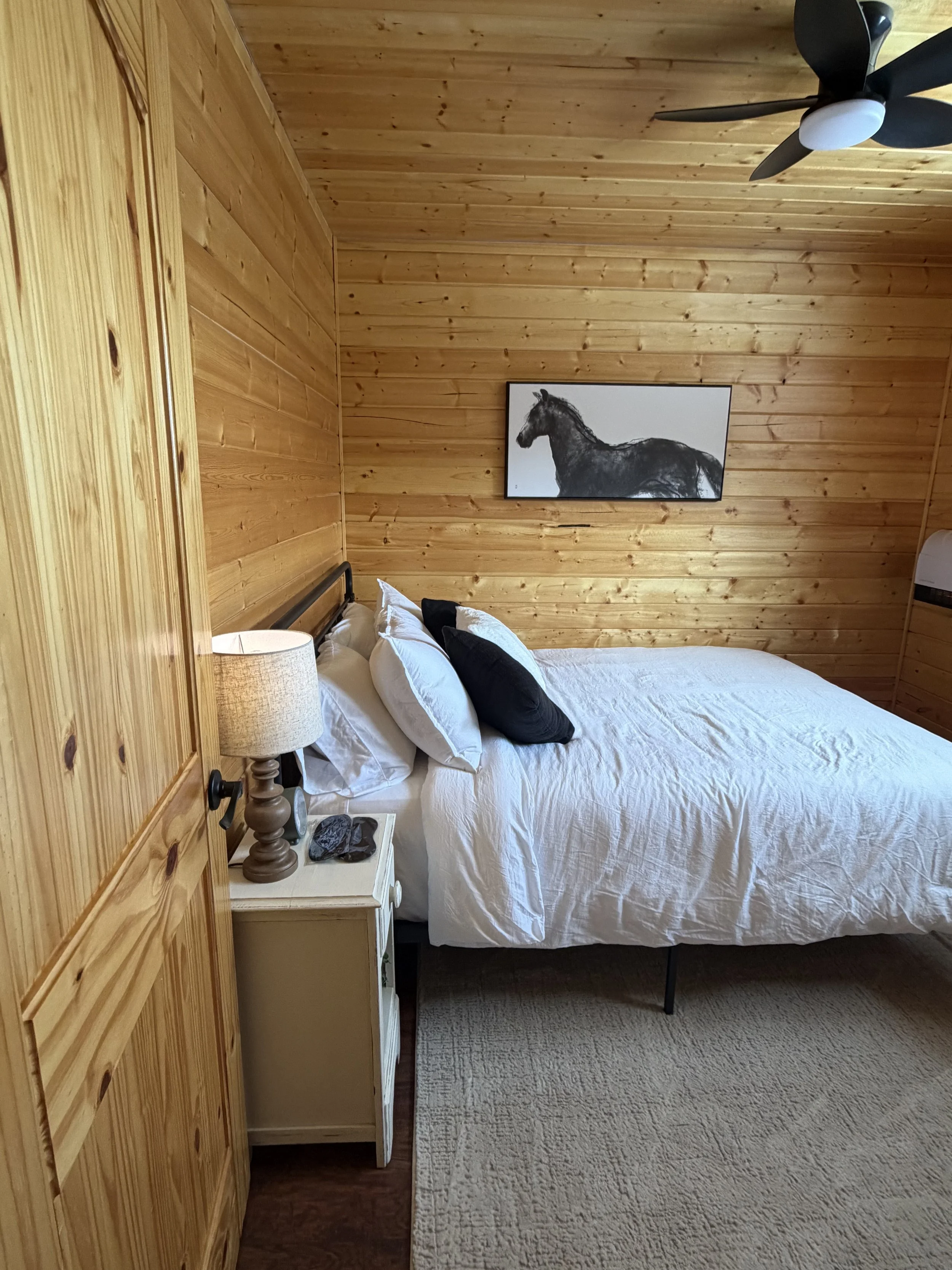A cozy bedroom with wood-paneled walls, a white bed with black and white pillows, a nightstand with a lamp, and a black and white horse painting on the wall.