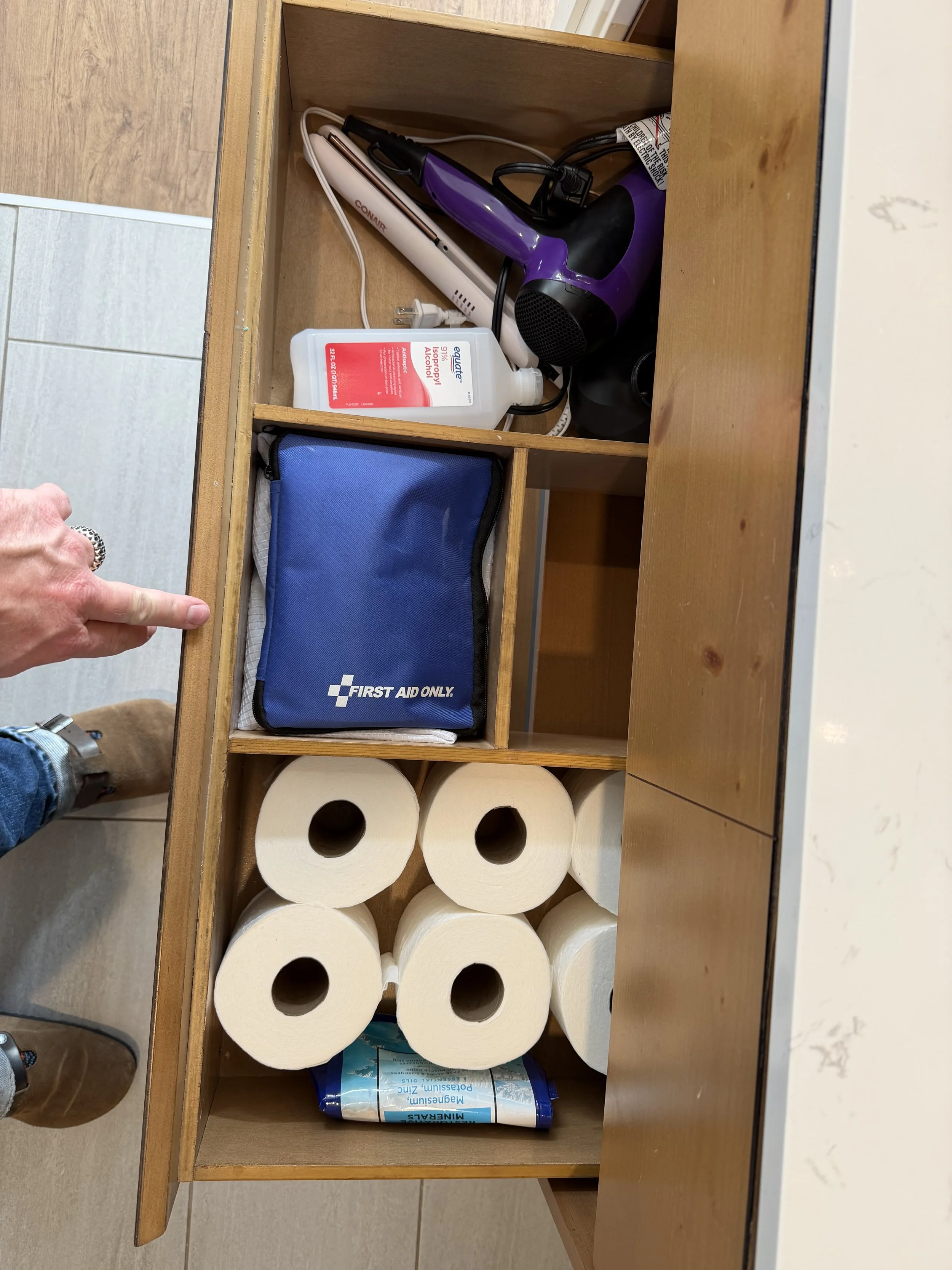 Open wooden cabinet drawer containing a blue first aid kit, six rolls of toilet paper, a bottle of alcohol-based hand sanitizer, a purple hairdryer, a white electric straightening iron, a hairbrush, and some cords.